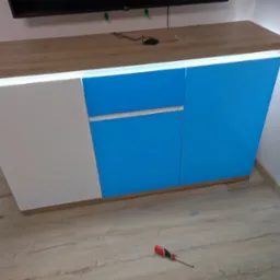 Nowoczesna szafka RTV z oświetleniem LED, biało-niebieskie fronty, blat w kolorze jasnego drewna, widoczny śrubokręt na podłodze z paneli.