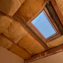 Poddasze z oknem dachowym w trakcie ocieplania, widoczna wełna mineralna między krokwiami, zabezpieczona sznurkami, ściana z widocznymi śladami po pracach remontowych.