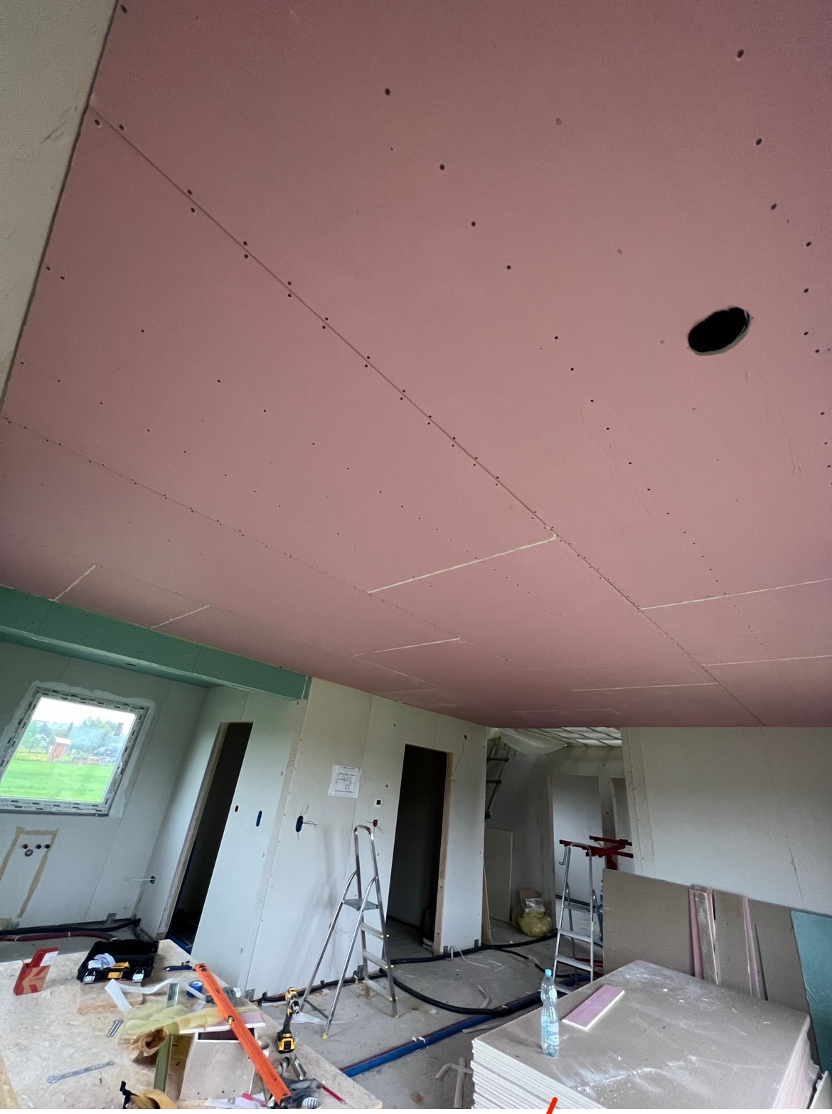 Różowa płyta karton gips na suficie w trakcie montażu, widoczne otwory na wkręty i otwór na oświetlenie, wnętrze w stanie surowym, drabiny i materiały budowlane w tle.