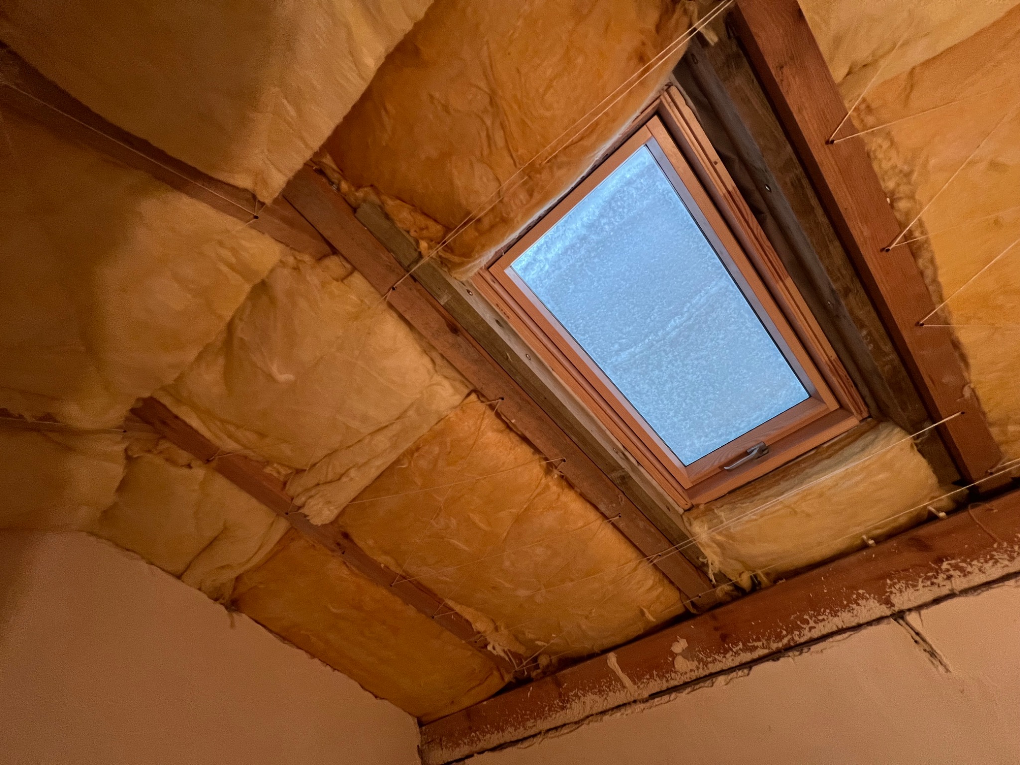 Poddasze z oknem dachowym w trakcie ocieplania, widoczna wełna mineralna między krokwiami, zabezpieczona sznurkami, ściana z widocznymi śladami po pracach remontowych.