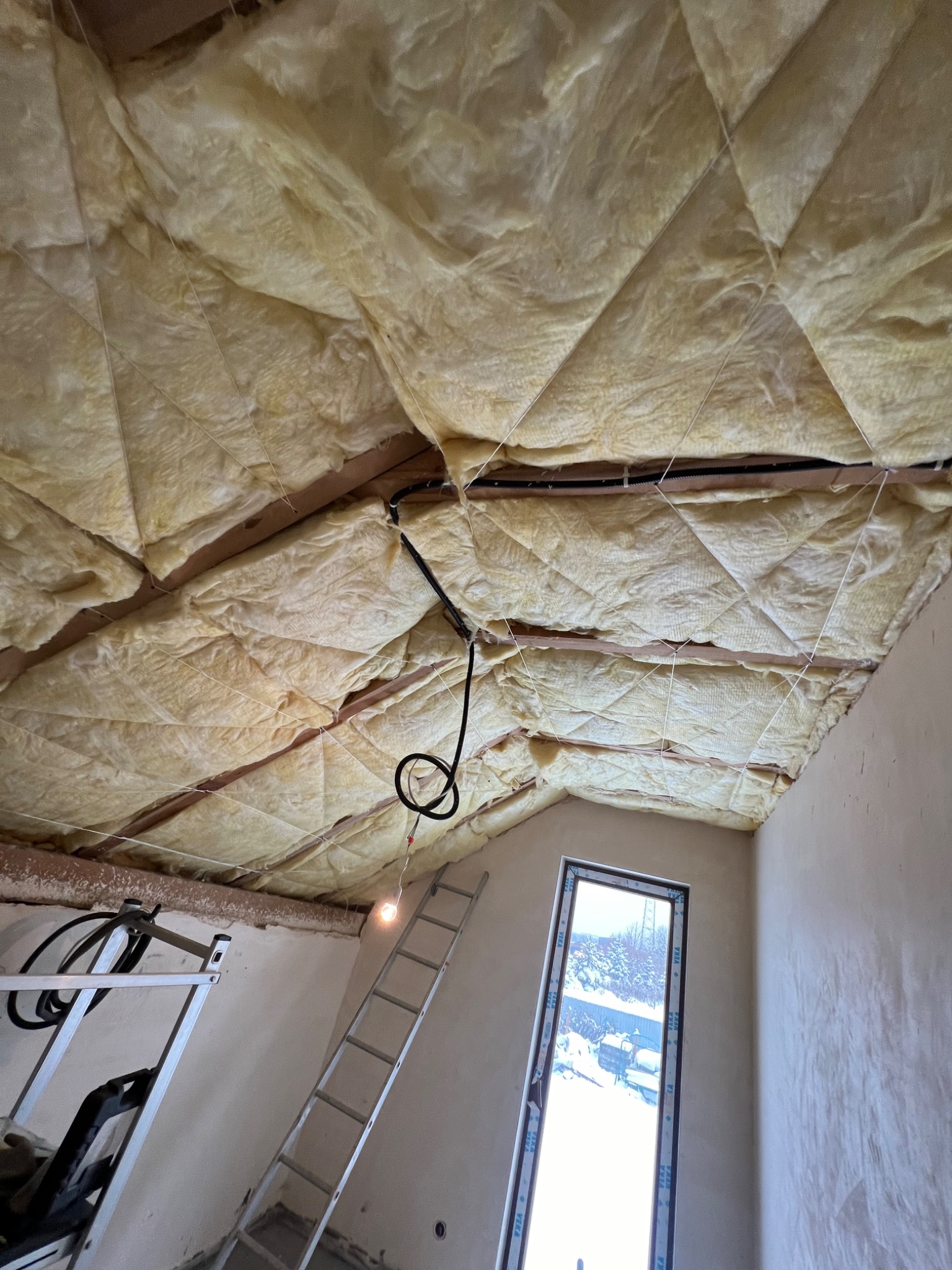 Izolacja poddasza wełną mineralną, widoczne drewniane belki, czarne kable elektryczne, drabina i okno z widokiem na śnieg.