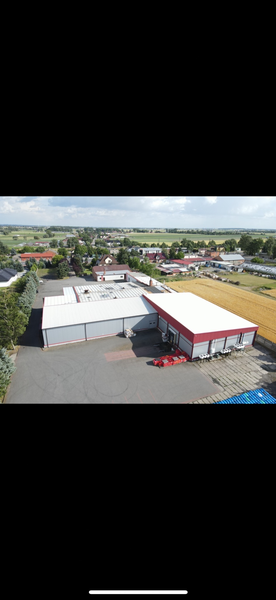 Widok z lotu ptaka na magazyn z rampą załadunkową, stertą czerwonych skrzynek i niebieskich beczek na zewnątrz, w tle zabudowania wiejskie i pole zboża.