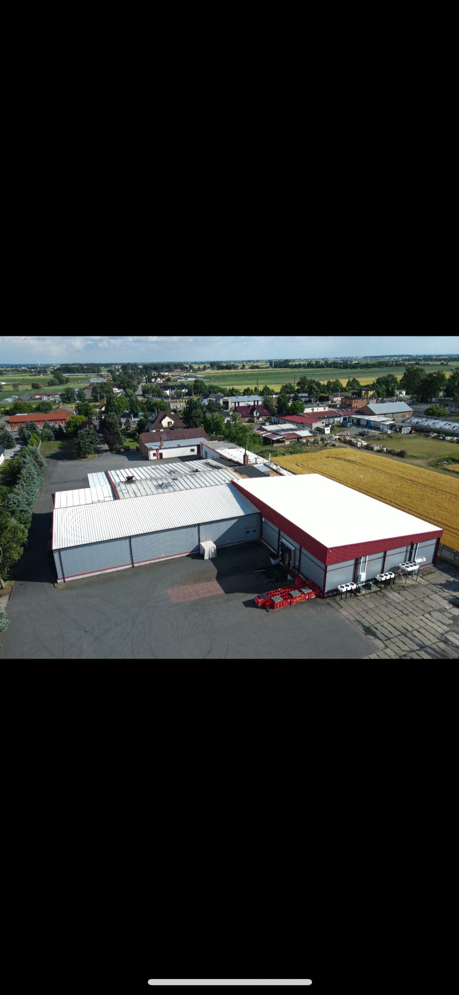 Zdjęcie z drona przedstawia budynek magazynowy z czerwonym dachem i szarymi ścianami, na zewnątrz stoją puste czerwone skrzynki, w tle widok na wieś.