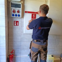 Bezpieczeństwo Ogniowe - usługi przeciwpożarowe Jakub Mrozowski i Wiktor Biernacki spółka cywilna - Instalator w Lublinie poziomuje tablicę z oznaczeniem przeciwpożarowym, widoczna gaśnica i skrzynka elektryczna.
