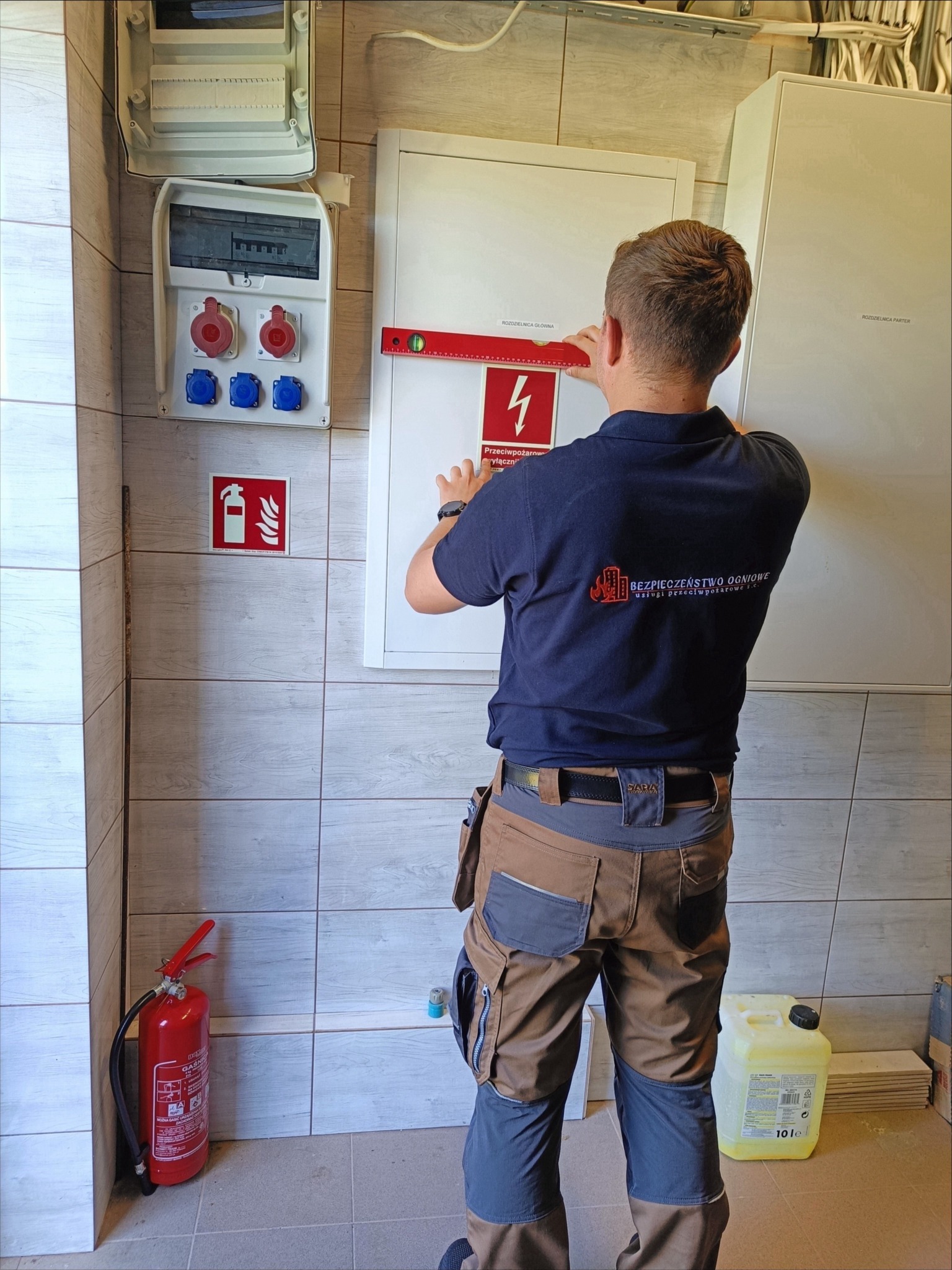 Instalator w Lublinie poziomuje tablicę z oznaczeniem przeciwpożarowym, widoczna gaśnica i skrzynka elektryczna.