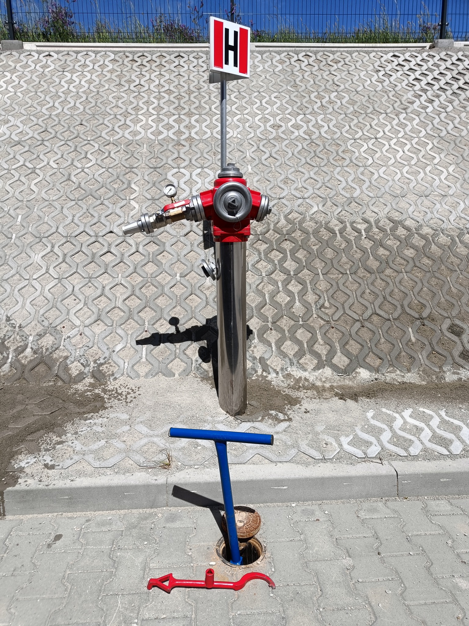 Czerwony hydrant zewnętrzny z manometrem i oznaczeniem 'H' na tle betonowej ściany z ażurowych bloków. Obok leżą narzędzia do otwierania studzienki hydrantowej: niebieski klucz i czerwony hak.