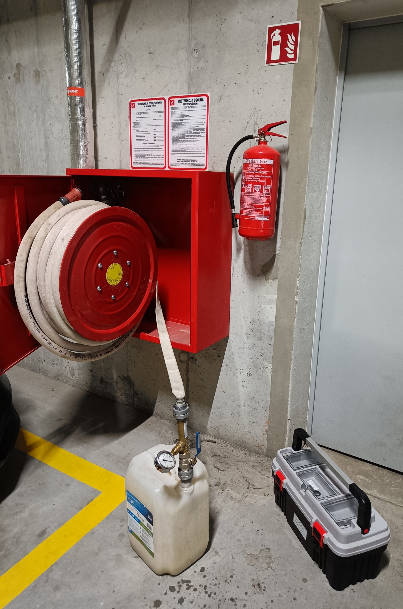 Czerwona szafka hydrantowa z rozwiniętym wężem, gaśnica, instrukcje ppoż, pojemnik z manometrem i skrzynka narzędziowa na tle betonowej ściany w garażu podziemnym.
