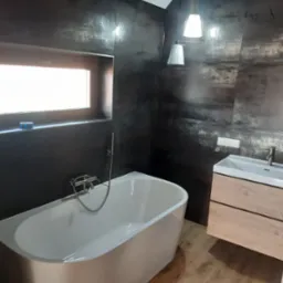 Nowoczesna łazienka w Szczytnie z wolnostojącą białą wanną, umywalką z szafką z drewnopodobnego materiału i ciemnymi płytkami imitującymi metal na ścianach.