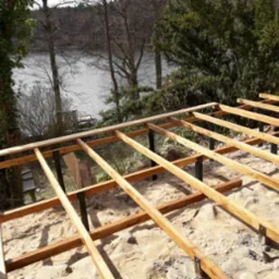 Drewniana konstrukcja pod podłogę tarasu na metalowych słupkach, widok na jezioro i drzewa w tle.