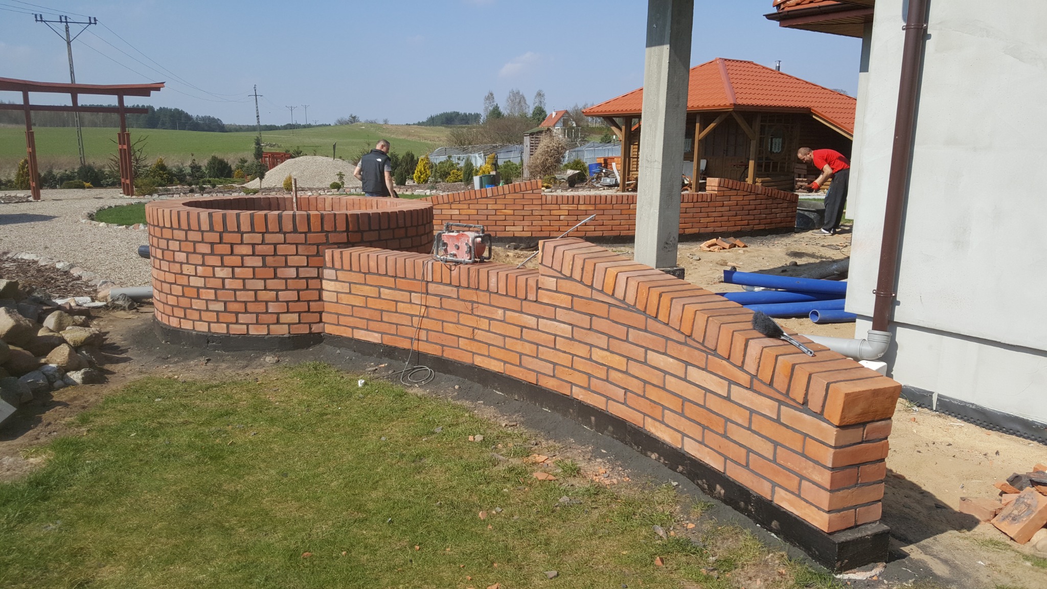 Budowa muru z czerwonej cegły z ciemną fugą, widoczny okrągły element i fragment budynku w tle, dwóch pracowników przy pracy.