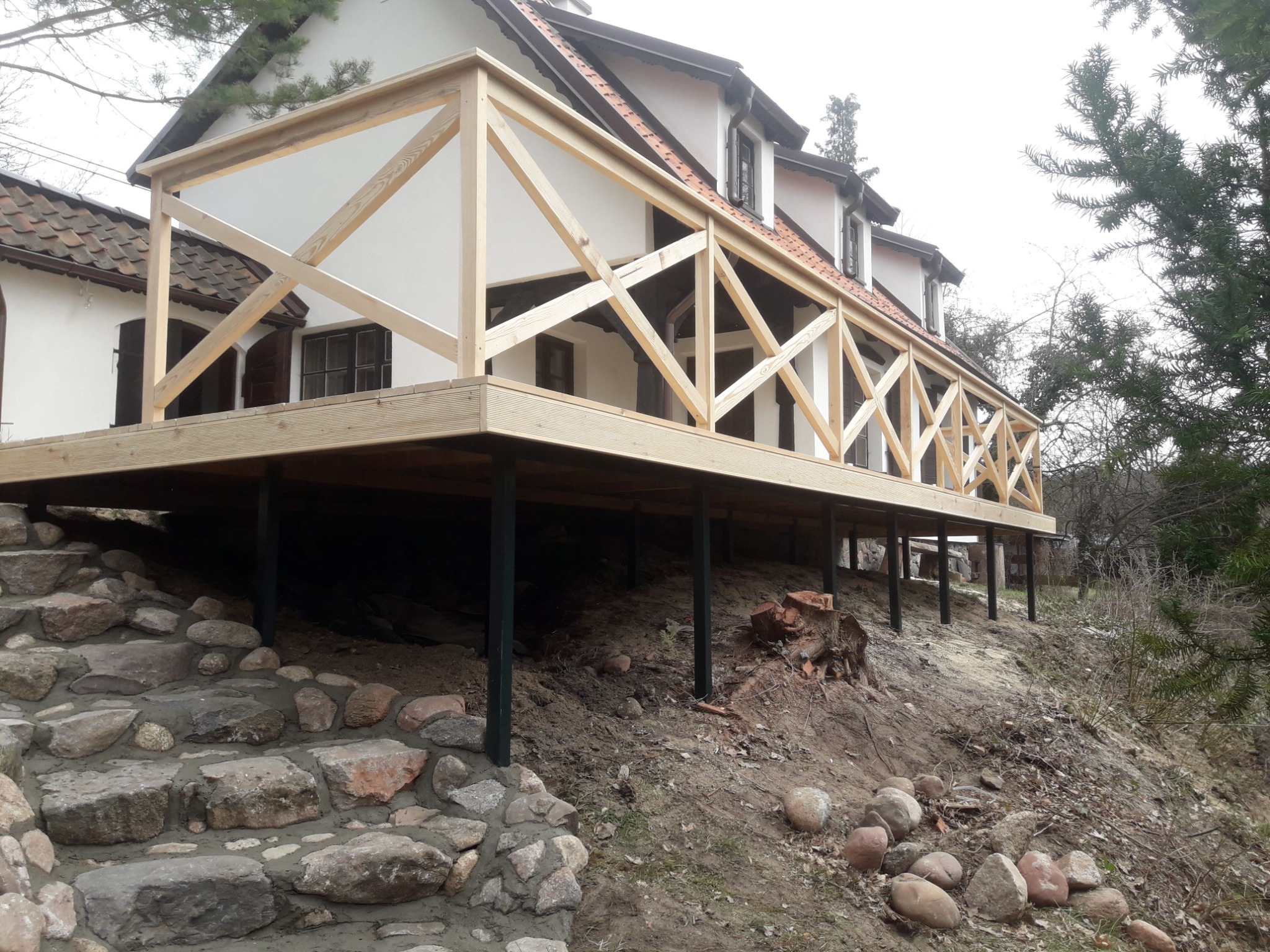 Nowo wybudowany drewniany taras z balustradą przy białym, dwupiętrowym domu z brązowym dachem, osadzony na czarnych metalowych słupach, z kamiennymi schodami prowadzącymi do ogrodu.