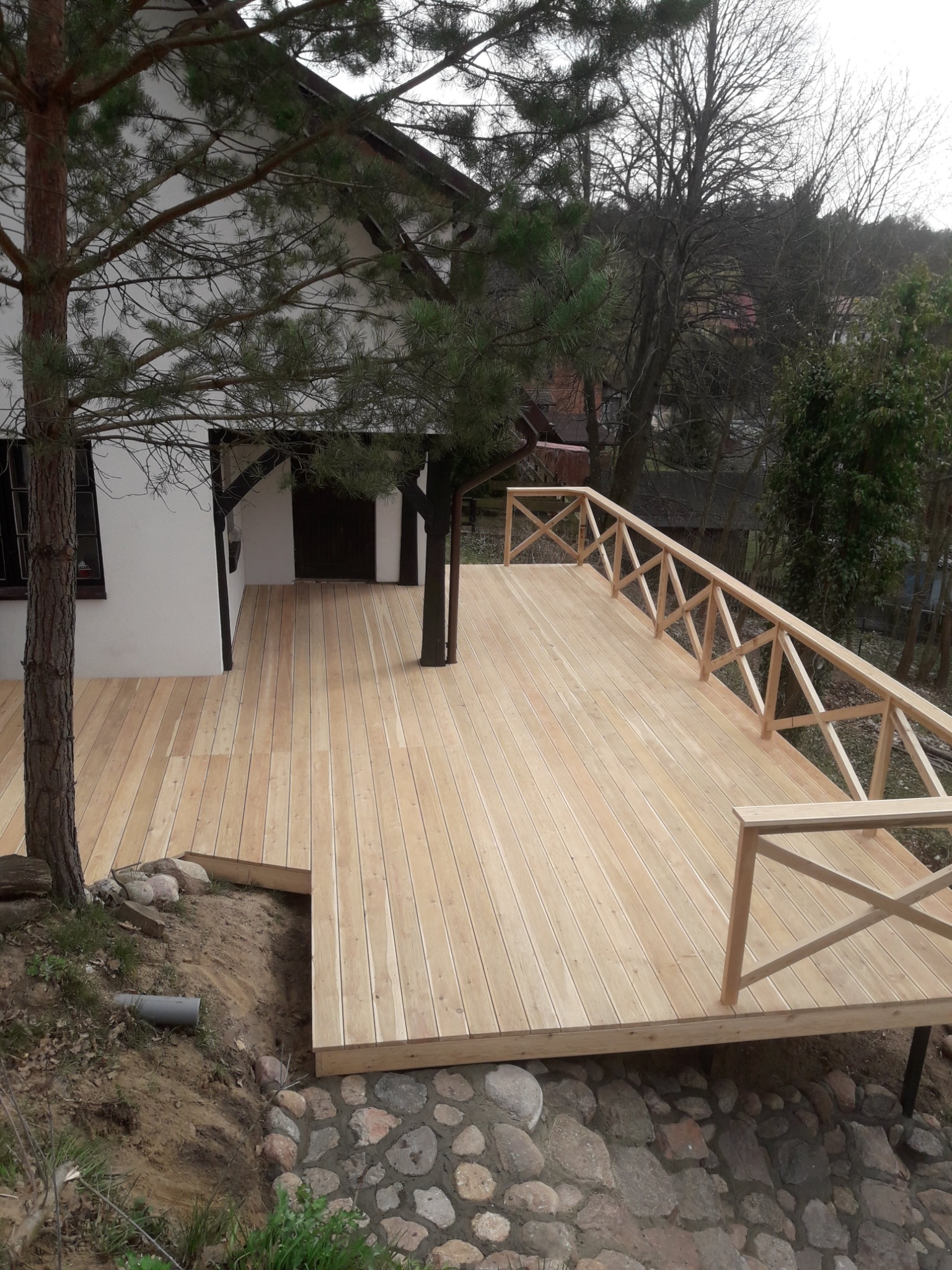 Nowo wybudowany drewniany taras z balustradą przy białym domu z drzewem rosnącym przez środek konstrukcji.