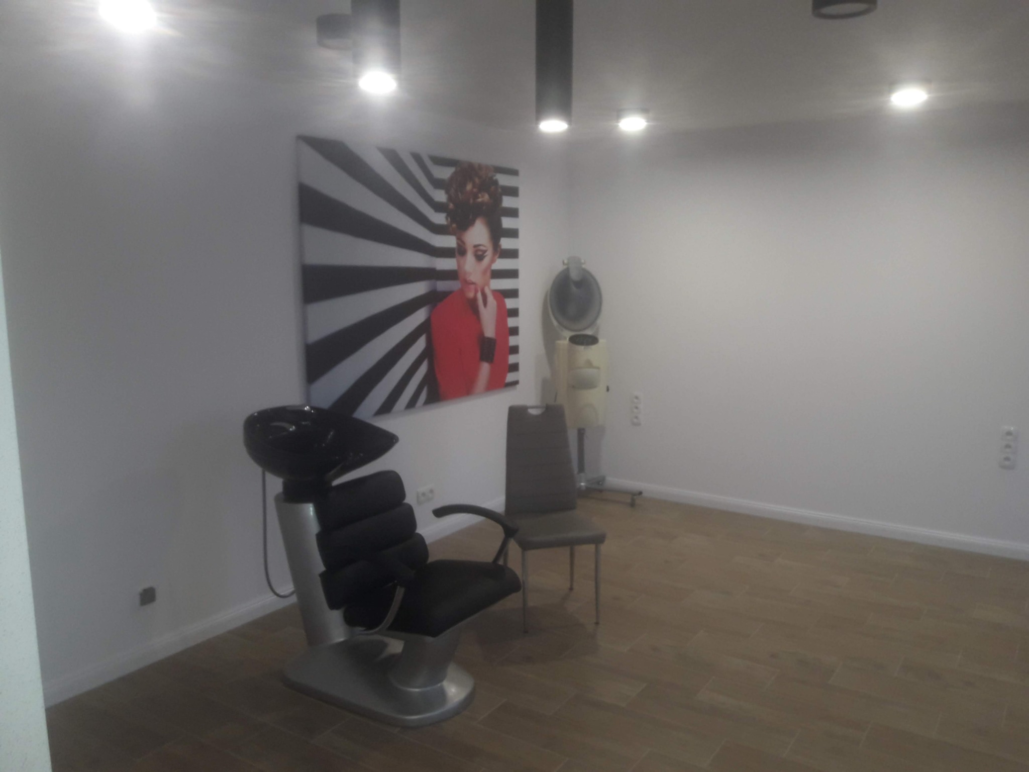 Wnętrze salonu fryzjerskiego z fotelem do mycia włosów, krzesłem i suszarką hełmową, na ścianie dekoracyjny obraz z motywem kobiety w geometrycznym tle.