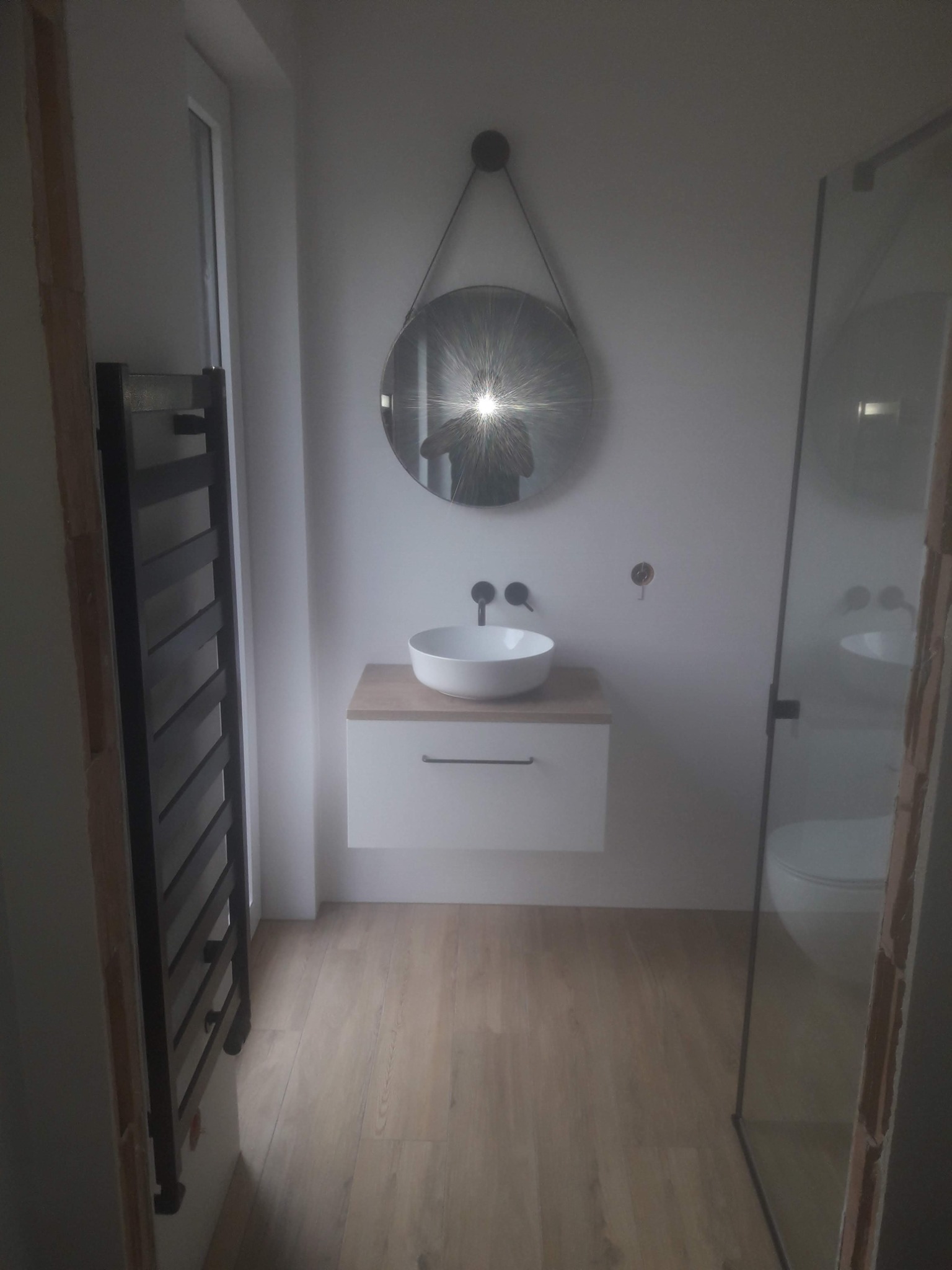 Nowoczesna łazienka z okrągłym lustrem zawieszonym na sznurku, białą umywalką na szafce z drewnianym blatem, grzejnikiem drabinkowym i kabiną prysznicową ze szklanymi drzwiami.