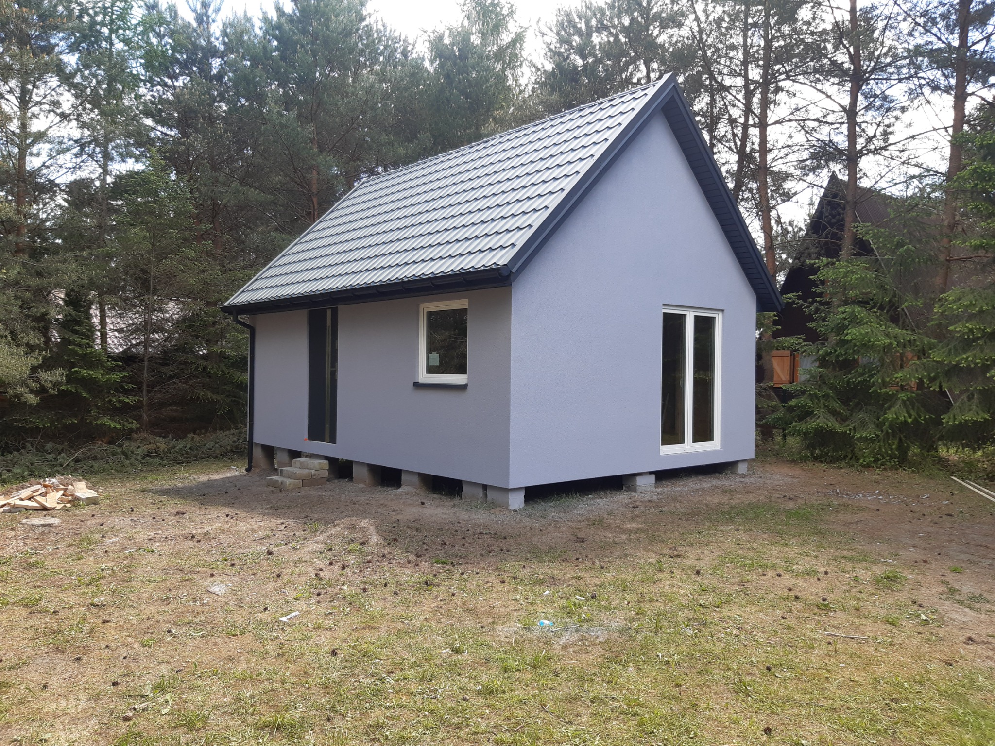 Niewielki, parterowy budynek z szarą elewacją i metalową dachówką, posadowiony na betonowych bloczkach w otoczeniu drzew iglastych.