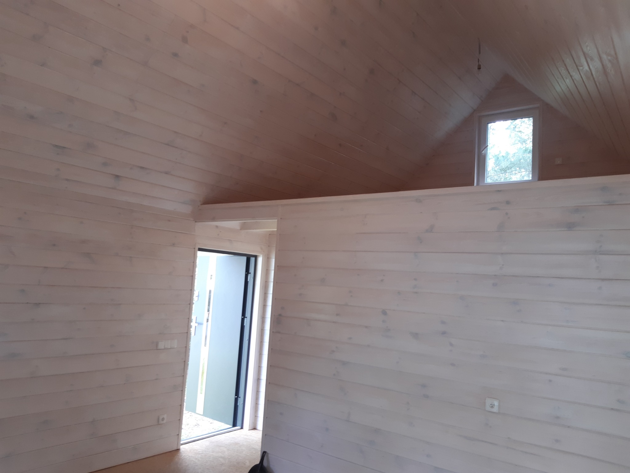 Wnętrze domu z boazerią pomalowaną na biało, widoczne skośne ściany poddasza, małe okno trójkątne w szczycie, drzwi wejściowe na wprost.