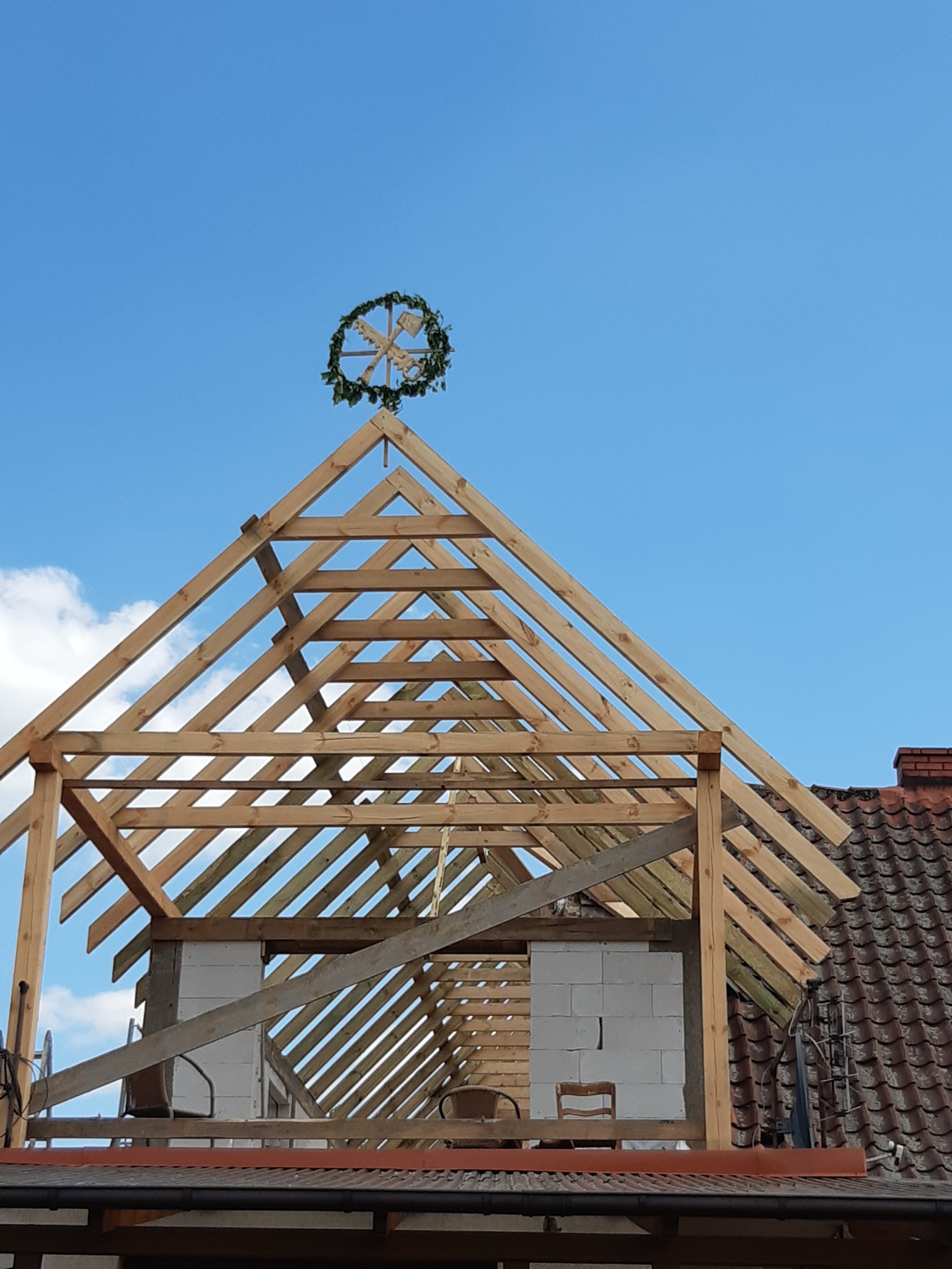 Wieniec ciesielski z narzędziami na szczycie drewnianej konstrukcji dachu domu w trakcie budowy z widocznymi bloczkami z betonu komórkowego, na tle błękitnego nieba.