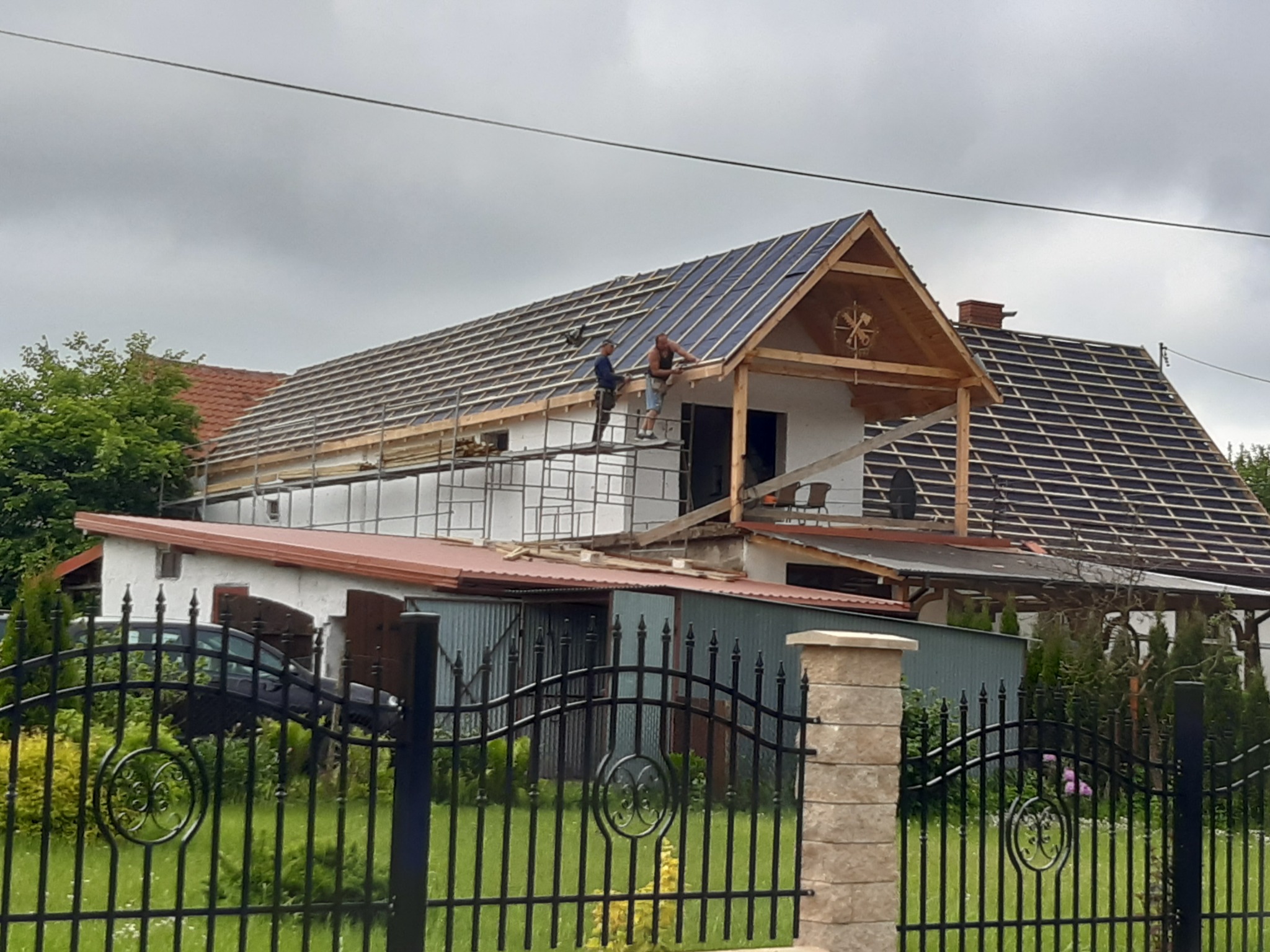 Dwóch mężczyzn na rusztowaniu pracuje nad konstrukcją dachu domu z drewnianymi elementami dekoracyjnymi na szczycie, widok zza czarnego metalowego ogrodzenia.