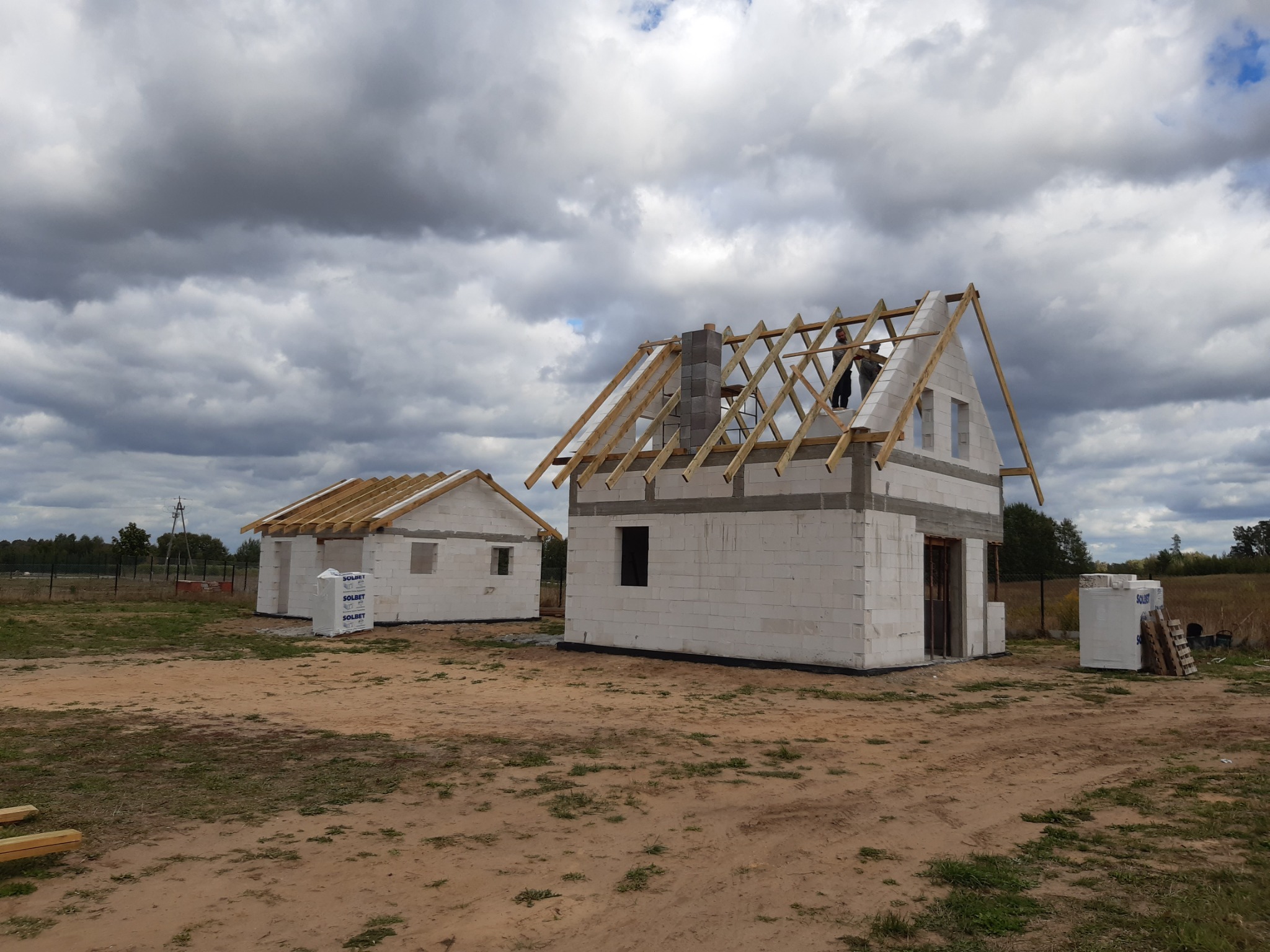 Dwa budynki w stanie surowym otwartym z białego bloczka, z drewnianą konstrukcją dachu, na działce z piaszczystą ziemią, pod pochmurnym niebem. Na drugim budynku widoczny komin z bloczków.