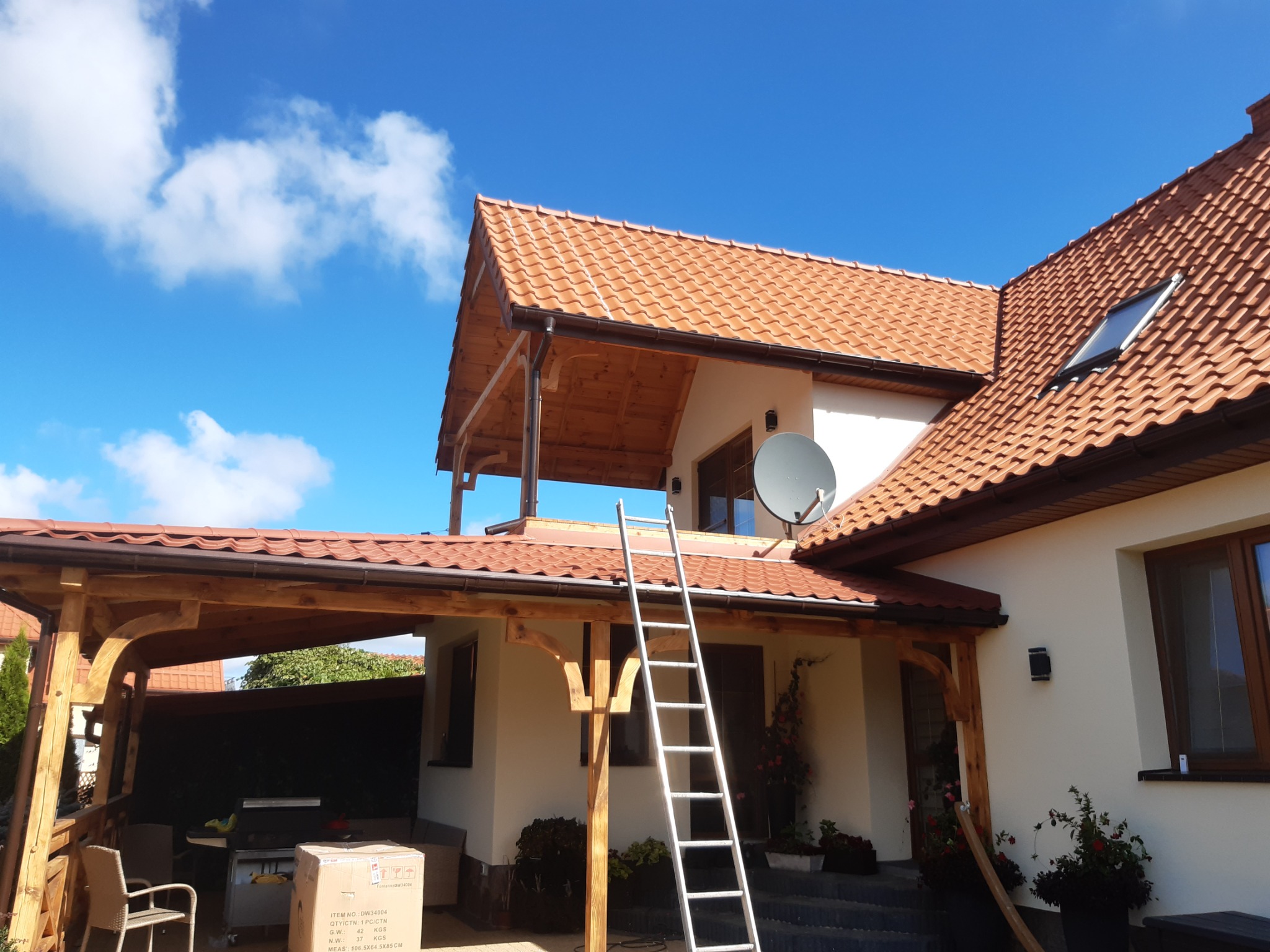 Nowoczesny dom murowany z czerwoną dachówką, drewnianą konstrukcją tarasu, anteną satelitarną na ścianie i drabiną opartą o ścianę, sfotografowany w słoneczny dzień z widocznymi chmurami.