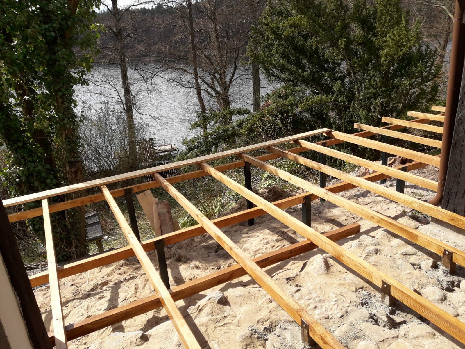 Drewniana konstrukcja pod podłogę tarasu na metalowych słupkach, widok na jezioro i drzewa w tle.