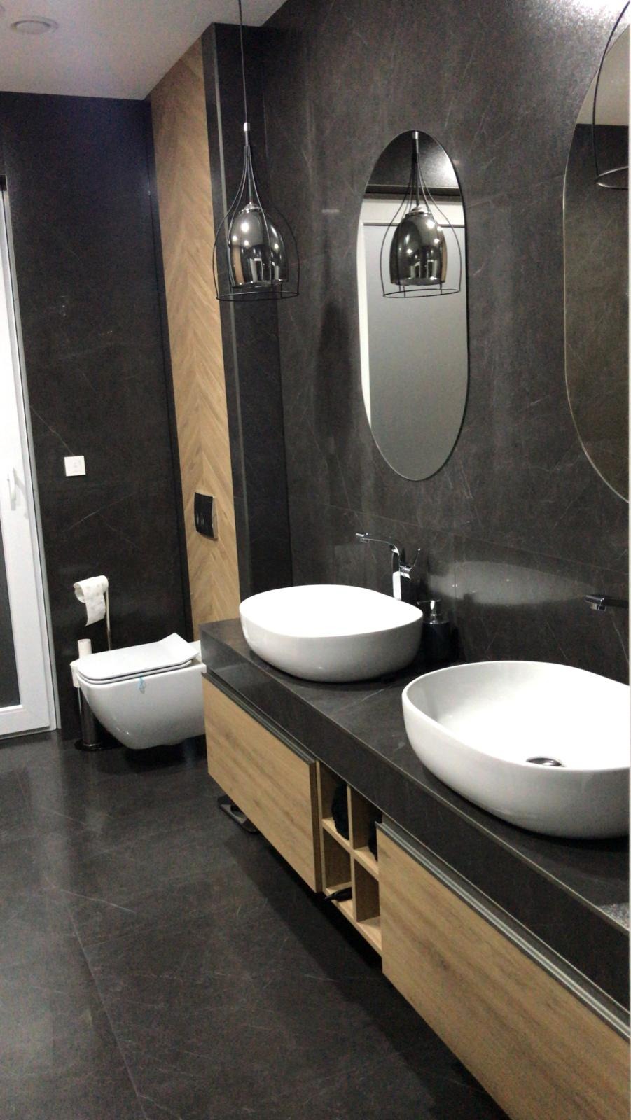 Nowoczesna łazienka z dwoma okrągłymi umywalkami nablatowymi, czarnymi płytkami imitującymi kamień na ścianach i podłodze, drewnianą szafką pod umywalkami oraz wiszącą toaletą z boku.