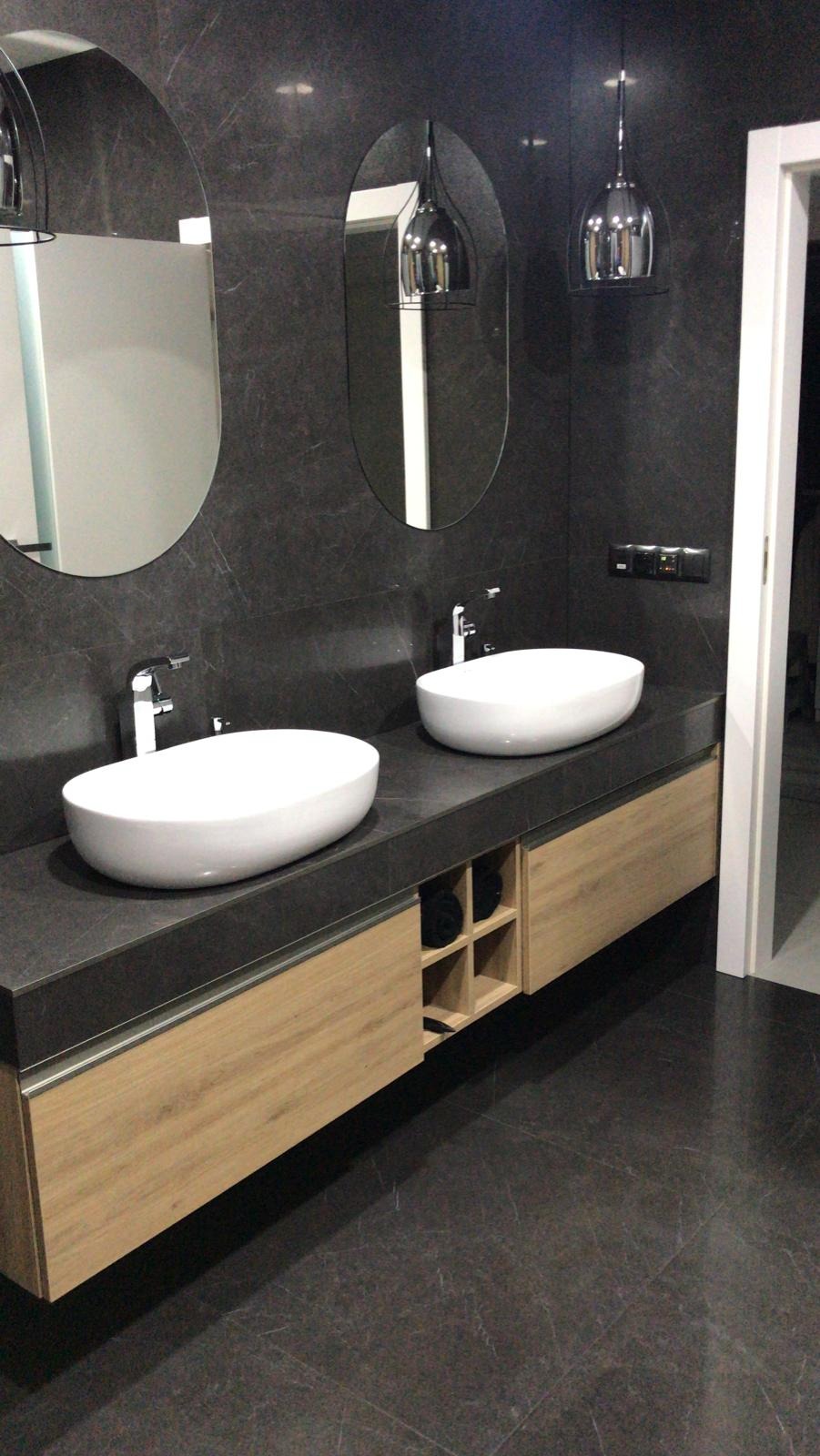 Nowoczesna łazienka z dwoma owalnymi umywalkami nablatowymi, ciemnoszarym blatem i podwieszaną szafką z drewnianymi frontami, podświetlona wiszącymi lampami w chromowanych obudowach, z lustrami...