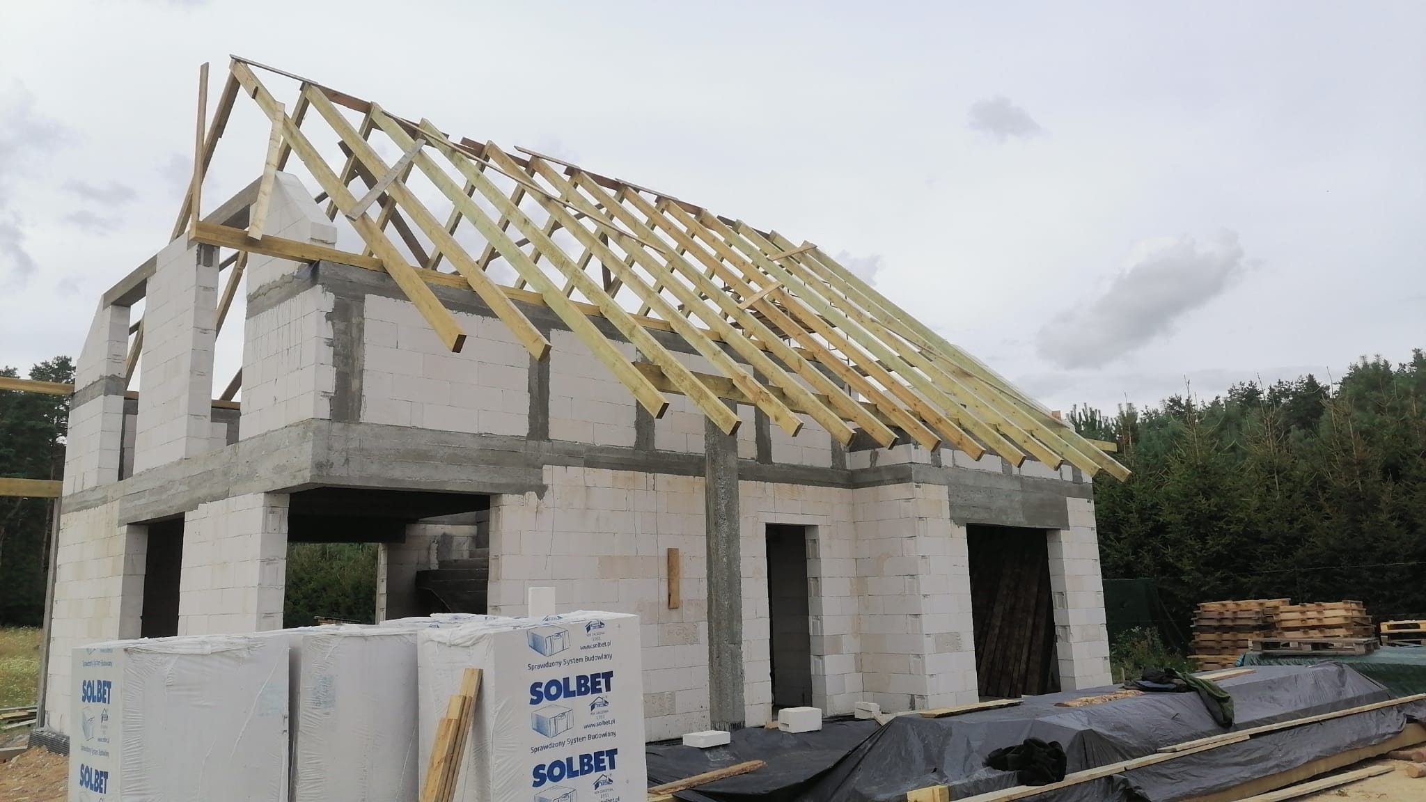 Wznoszony budynek z białych bloczków z widoczną konstrukcją drewnianą dachu, palety z bloczkami Solbet na pierwszym planie.