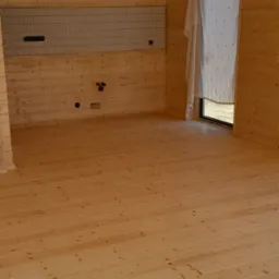 Surowe wnętrze drewnianego domu w trakcie wykończenia, widoczne deski podłogowe, ściany z boazerii, instalacje elektryczne i hydrauliczne w ścianach, okno zasłonięte tkaniną.