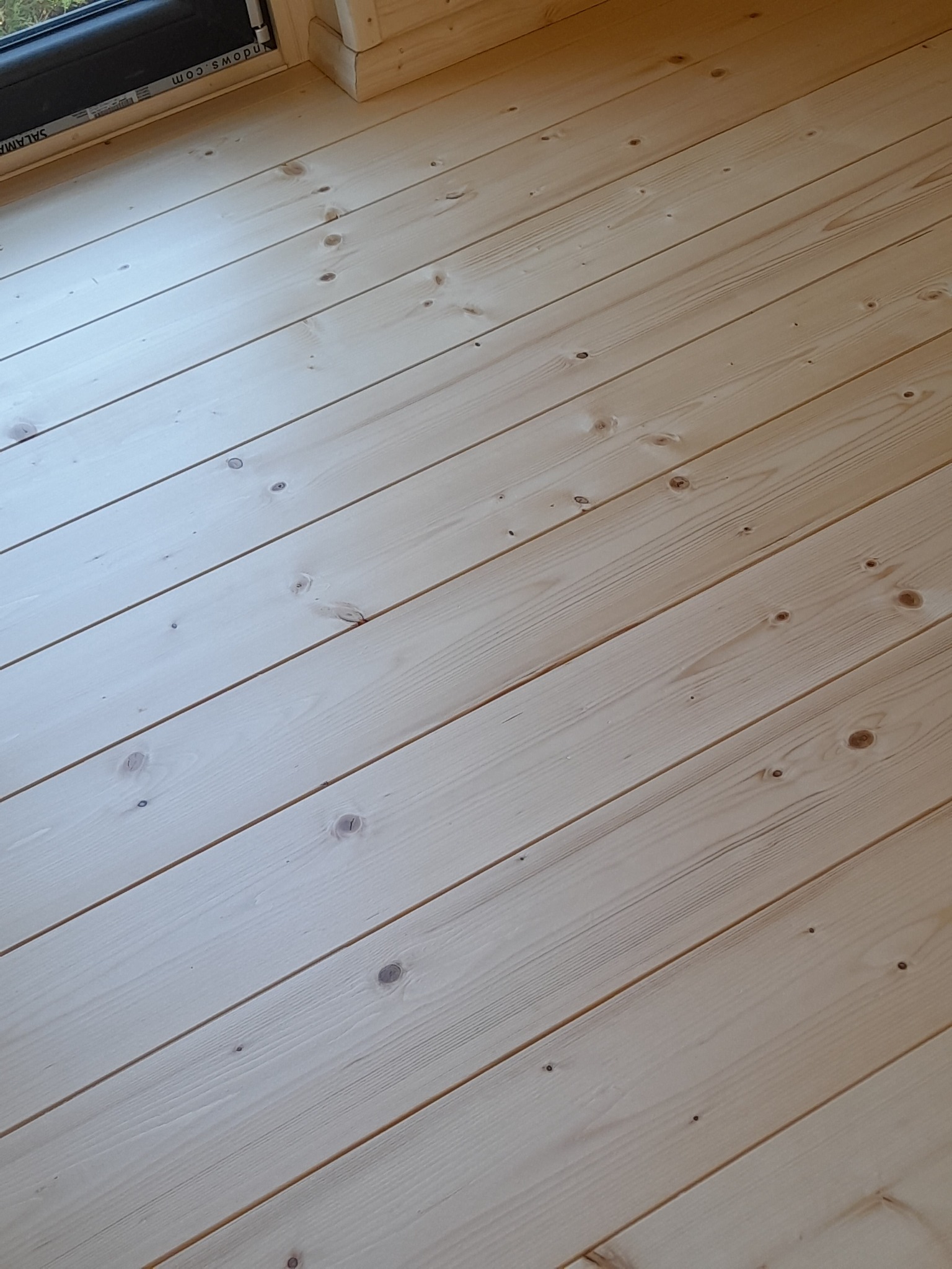 Jasna, sosnowa podłoga z widocznymi słojami, ułożona ukośnie, z fragmentem okna w lewym górnym rogu.