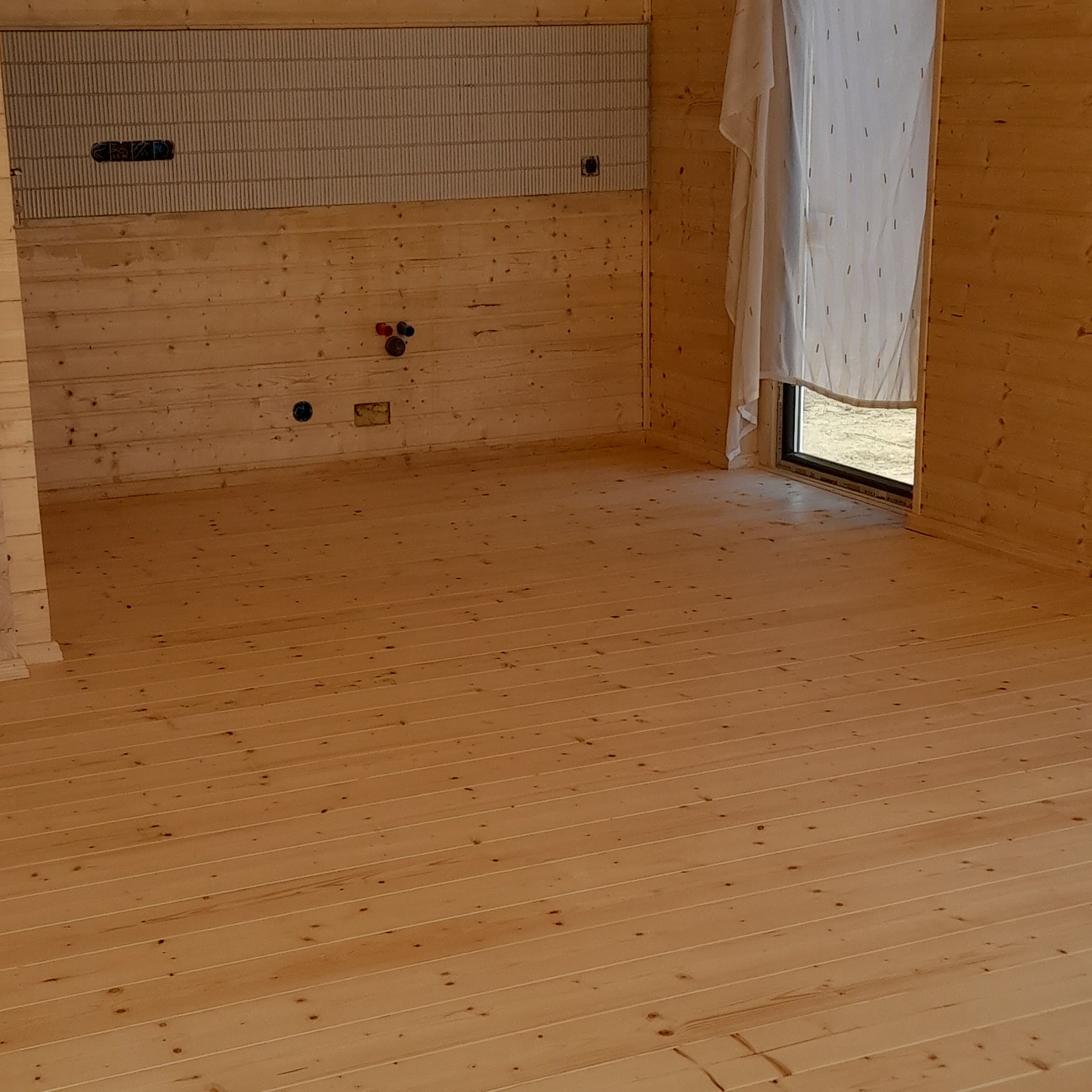 Surowe wnętrze drewnianego domu w trakcie wykończenia, widoczne deski podłogowe, ściany z boazerii, instalacje elektryczne i hydrauliczne w ścianach, okno zasłonięte tkaniną.