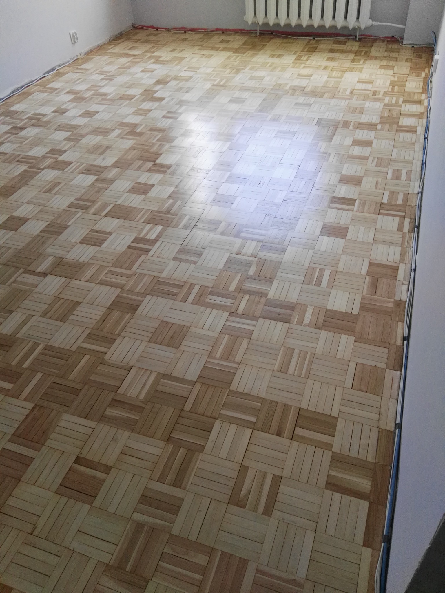 Ułożona parkietowa podłoga z drewnianych kwadratów w jasnych odcieniach brązu, widoczny fragment grzejnika i instalacji przy ścianie.