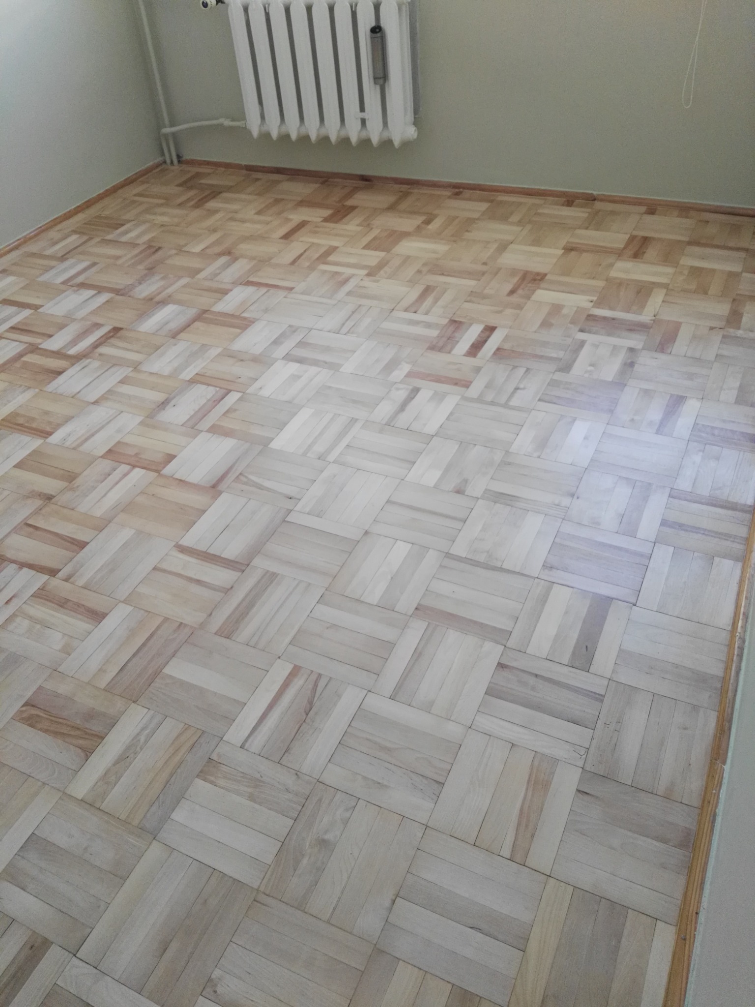 Parkiet ułożony w jodełkę z jasnego drewna, widoczny grzejnik na ścianie, neutralne oświetlenie.