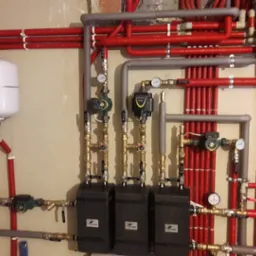 Złożony system rur grzewczych, z czerwonymi rurami z izolacją i szarymi rurami, z trzema czarnymi skrzynkami z logo, manometrami i zaworami, zamontowany na beżowej ścianie.