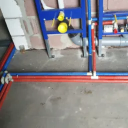 Instalacja wodno-kanalizacyjna w trakcie montażu: rury PEX w kolorach czerwonym i niebieskim ułożone wzdłuż ściany, widoczne stelaże podtynkowe z elementami przyłączeniowymi, surowy beton...