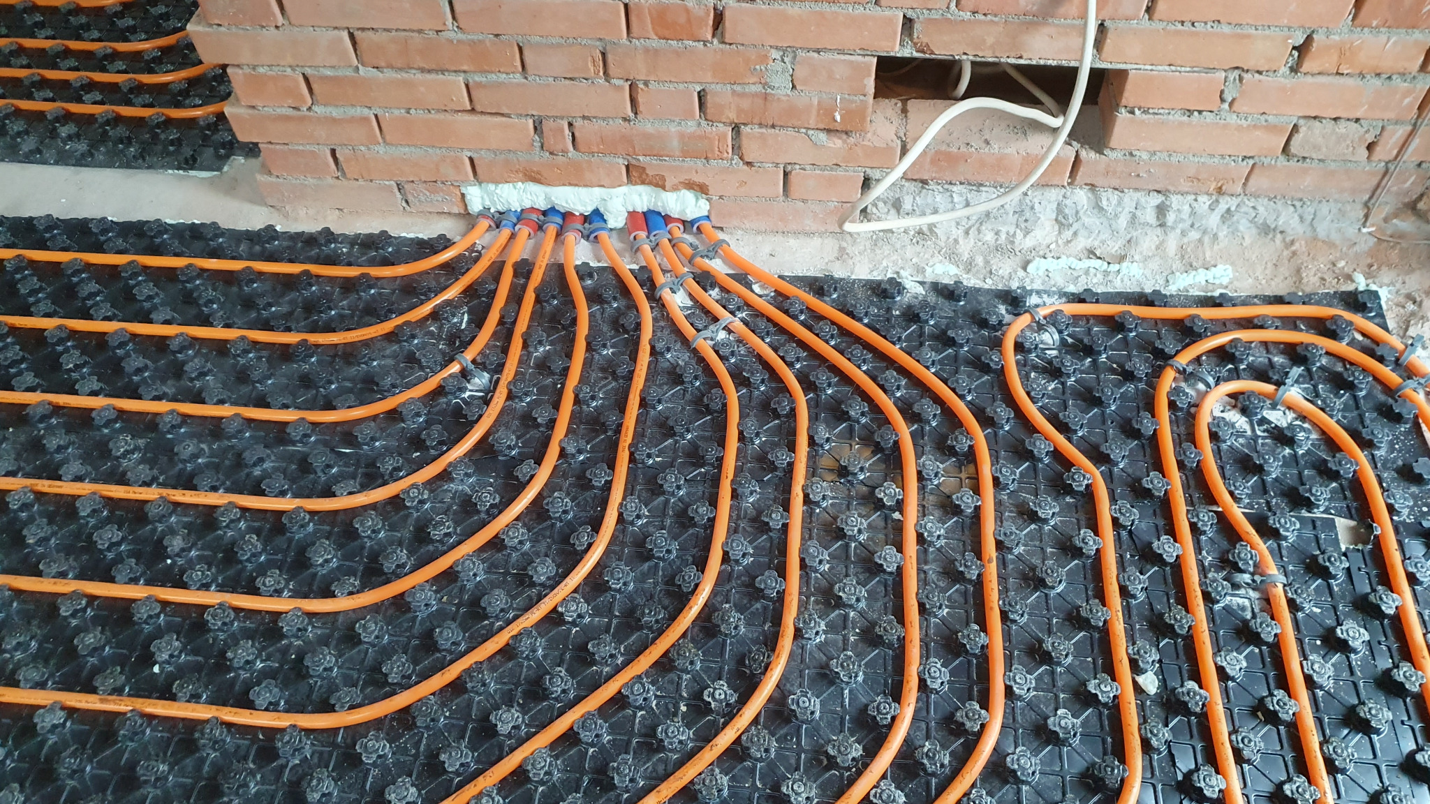 Pomarańczowe rury ogrzewania podłogowego ułożone na czarnej macie montażowej, prowadzące do ściany z czerwonej cegły z widoczną białą pianką uszczelniającą.