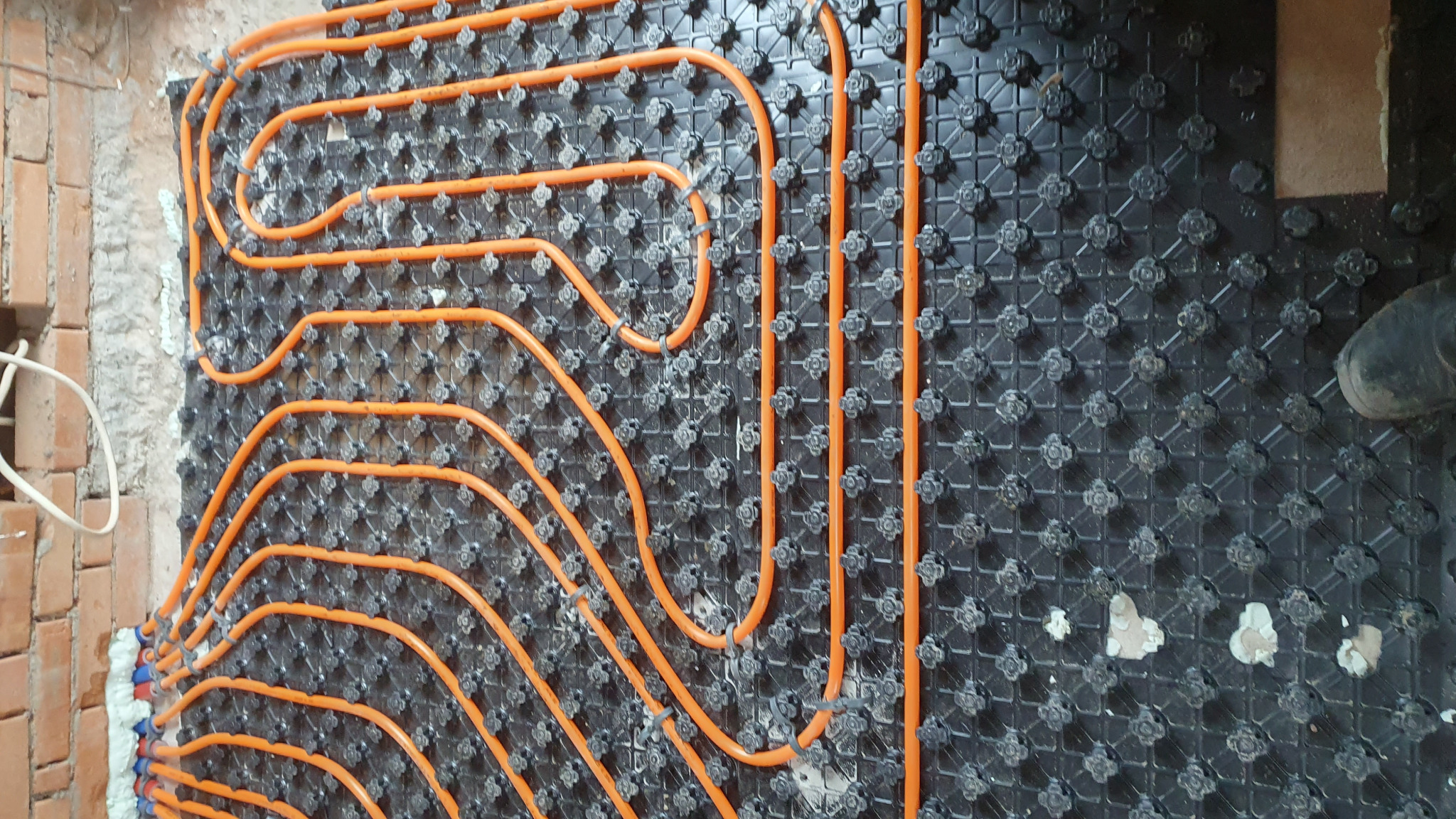 Pomarańczowe rury rozprowadzone na czarnej macie z wypustkami, przygotowane do zalania w systemie ogrzewania podłogowego, widoczne fragmenty muru z cegły.