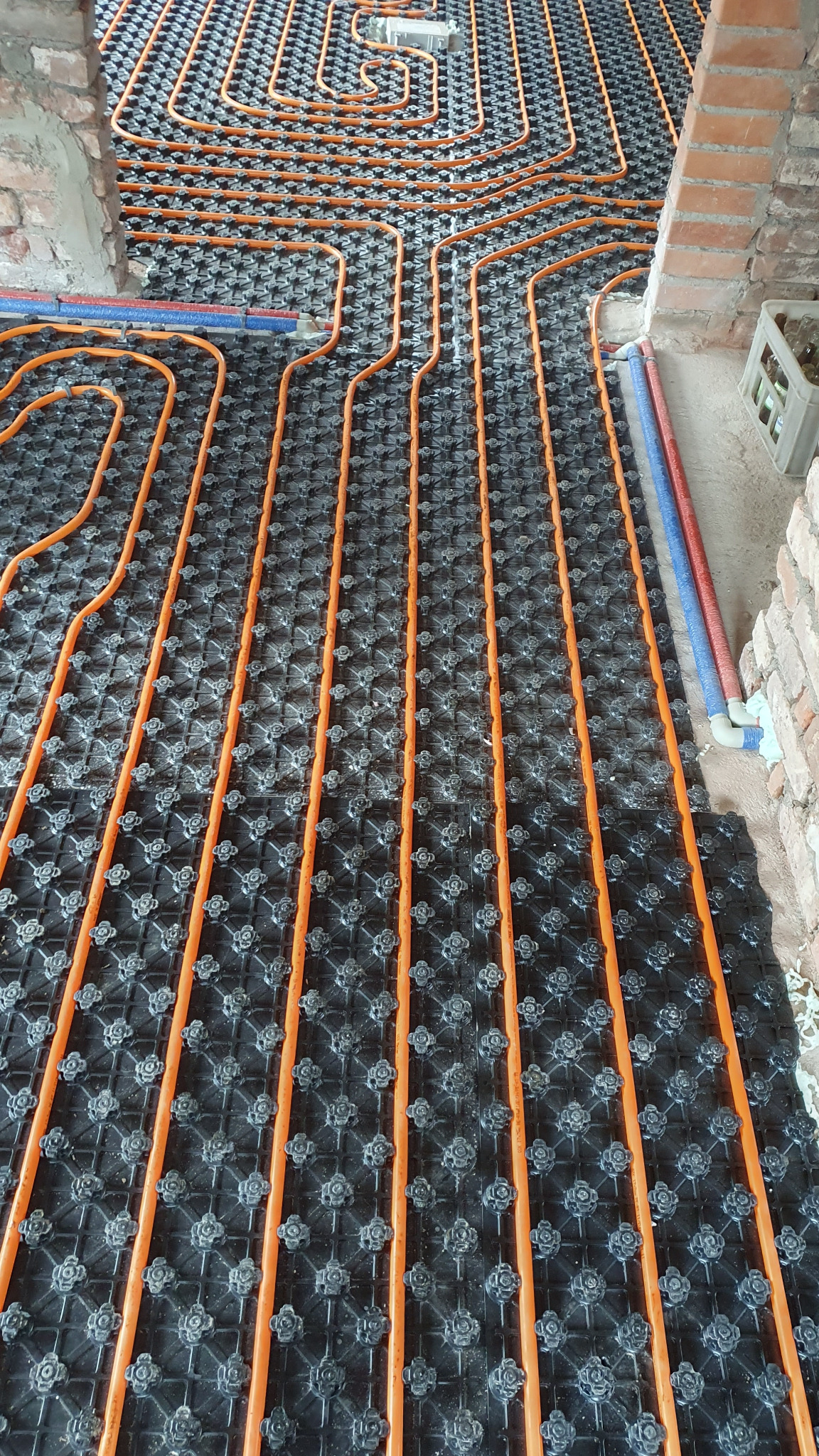 Pomarańczowe rury ogrzewania podłogowego rozłożone na czarnej macie systemowej, widoczne fragmenty muru z cegły i niebiesko-czerwone rury doprowadzające.