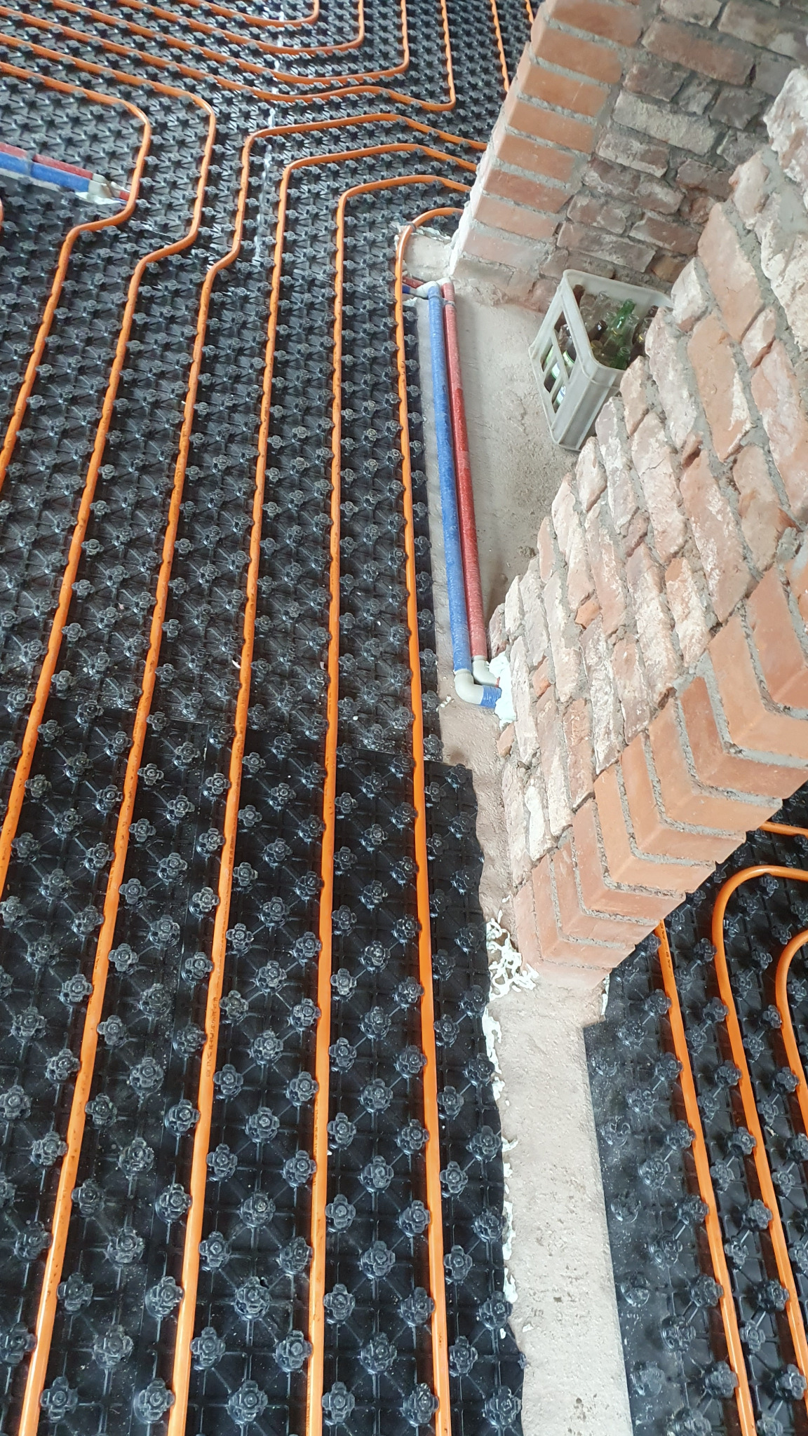 Pomarańczowe rurki ogrzewania podłogowego ułożone na czarnej macie z wypustkami, widoczne rury doprowadzające wodę oraz surowy, ceglany fragment ściany w tle.