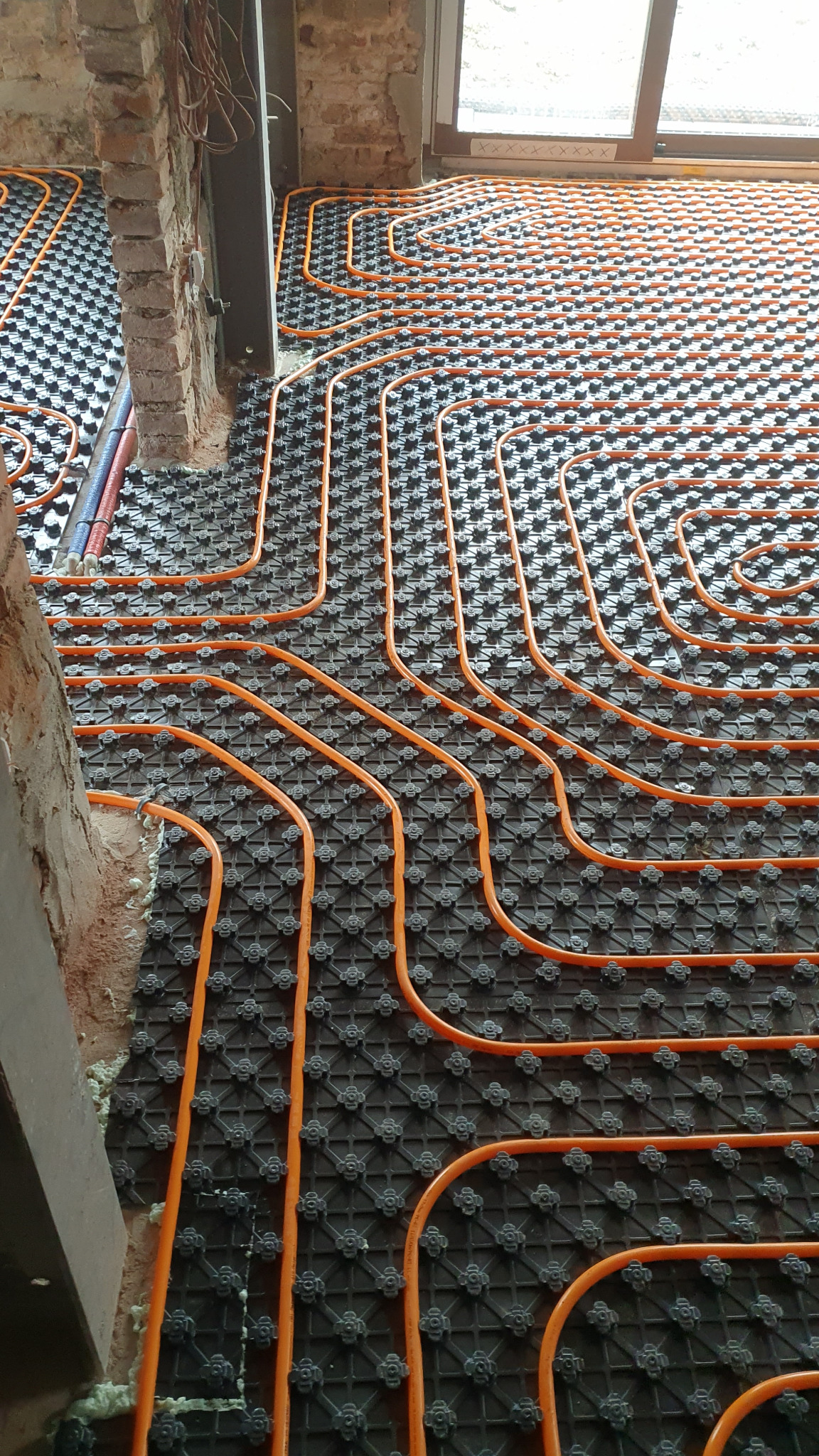 Pomarańczowe rury grzewcze ułożone na czarnej macie izolacyjnej z wypustkami, widok z góry na surową podłogę w trakcie prac instalacyjnych, fragment ceglanej ściany w tle.