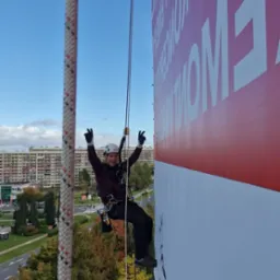 Pracownik na linie, w kasku, z podniesionymi rękami, na tle budynku i billboardu. Ujęcie z dołu, widok na miasto.