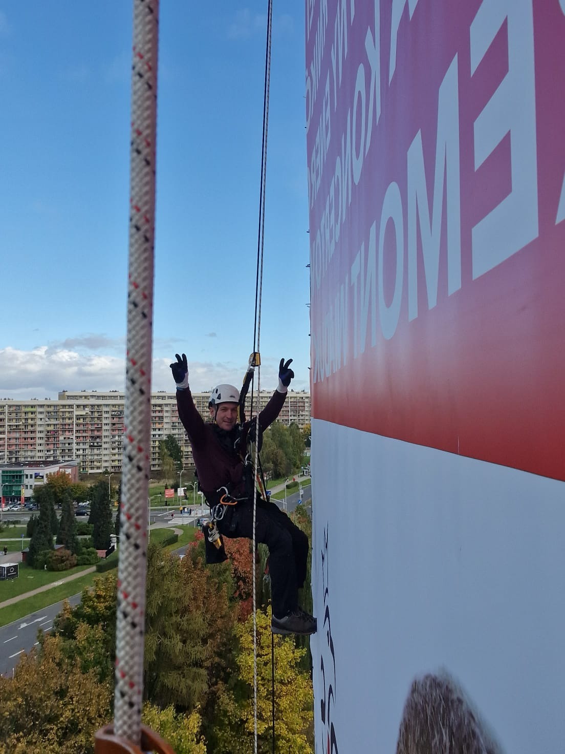 Pracownik na linie, w kasku, z podniesionymi rękami, na tle budynku i billboardu. Ujęcie z dołu, widok na miasto.
