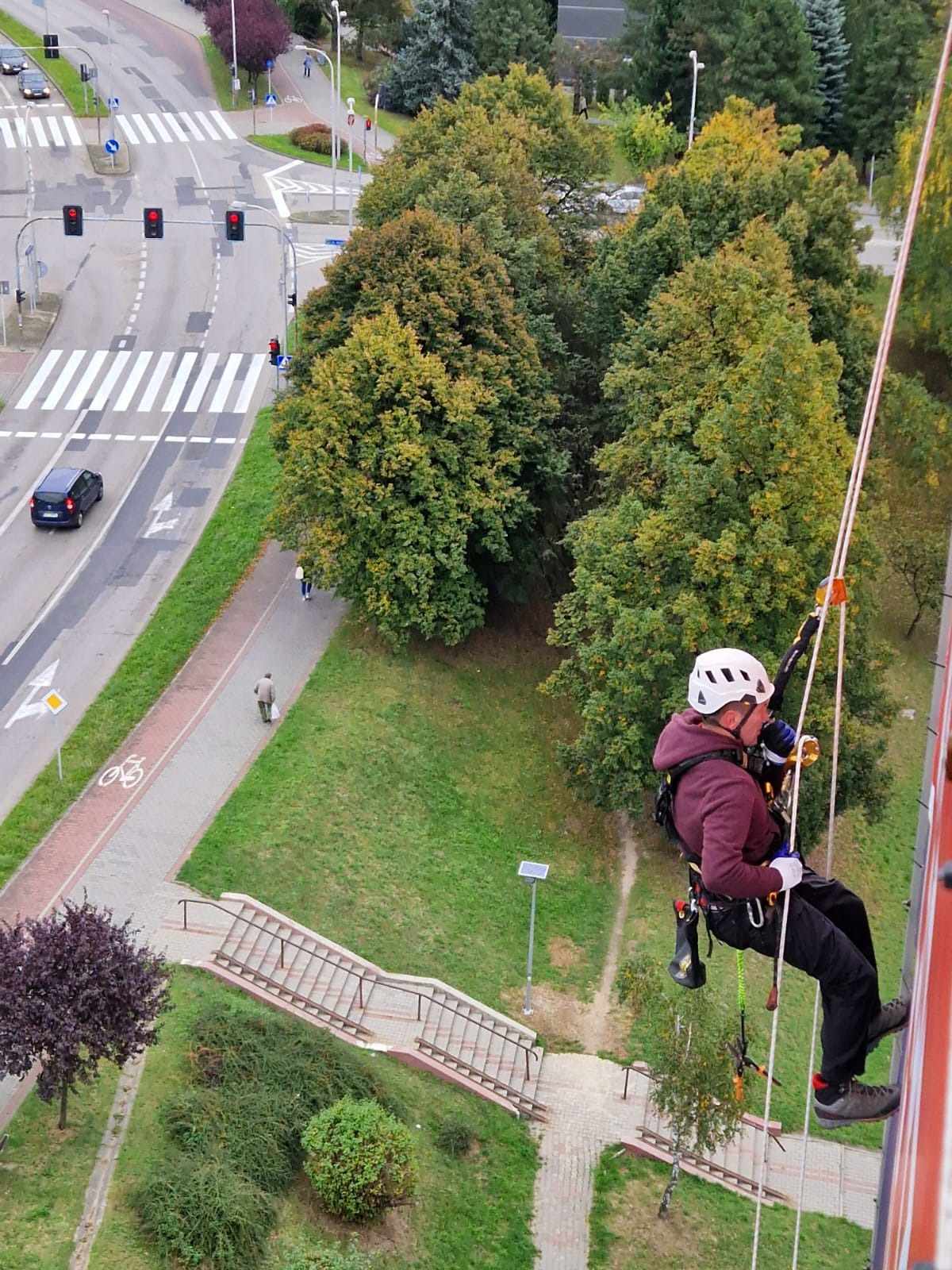 Pracownik w kasku, na linie, w trakcie pracy na wysokości, widok z góry na miasto z zielenią i ulicą.