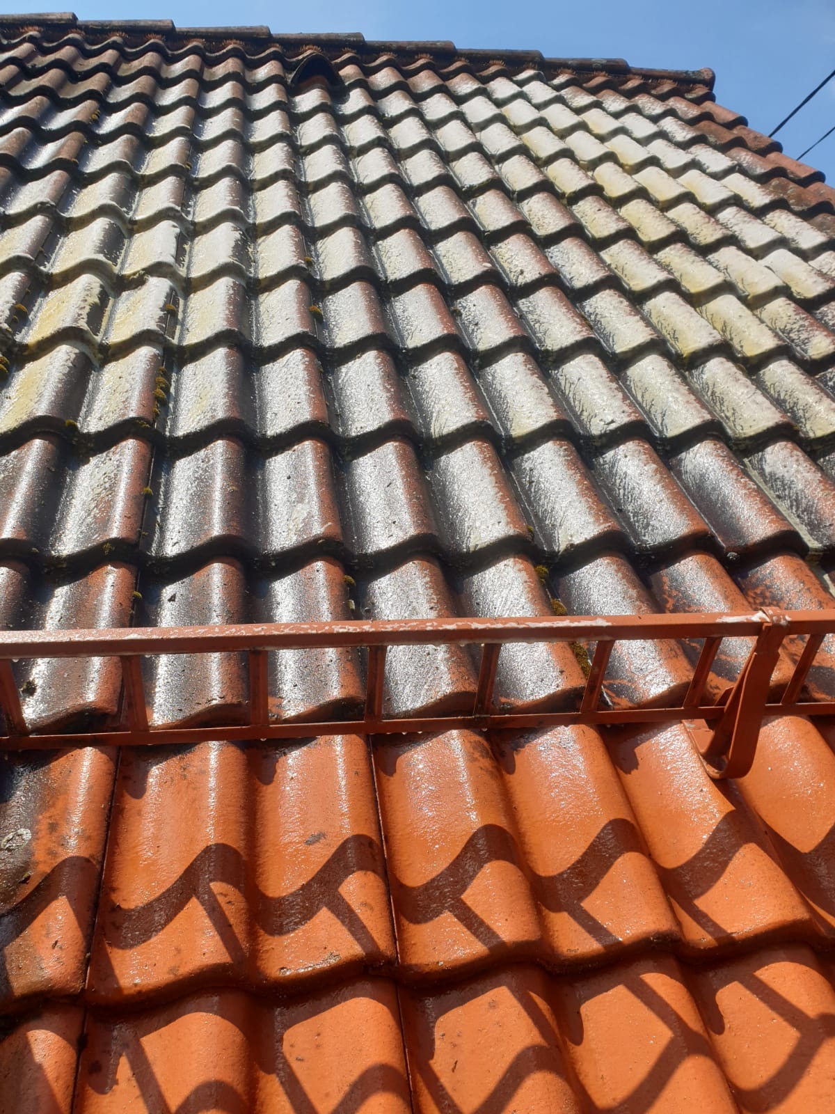 Czerwona, mokra dachówka po czyszczeniu, z resztkami mchu i metalowym zabezpieczeniem na dole kadru. Widok z dołu w słoneczny dzień.