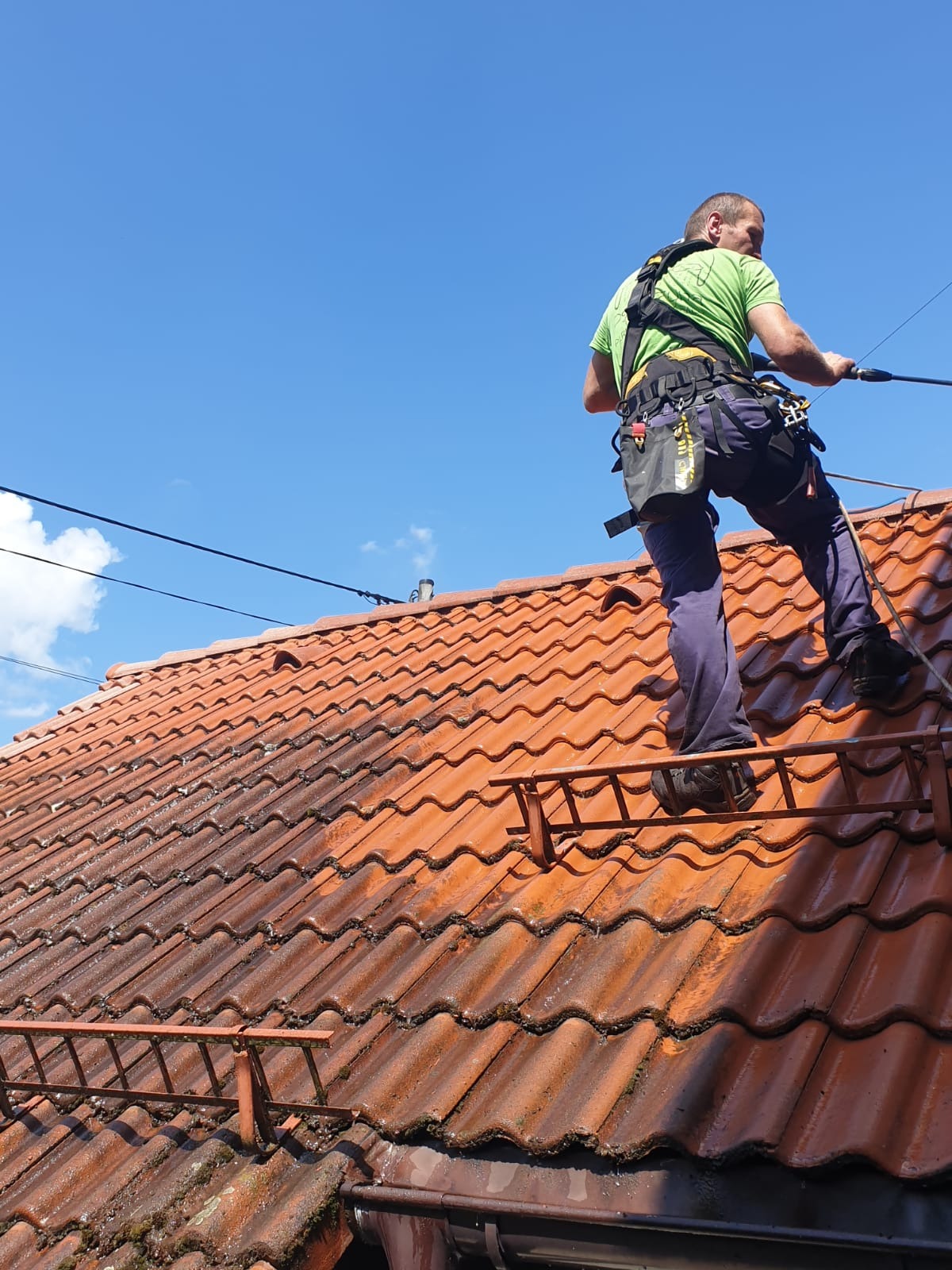 Pracownik w uprzęży na drabinie dachowej, czyści czerwoną dachówkę myjką ciśnieniową. Widoczna różnica czystości dachu, błękitne niebo.