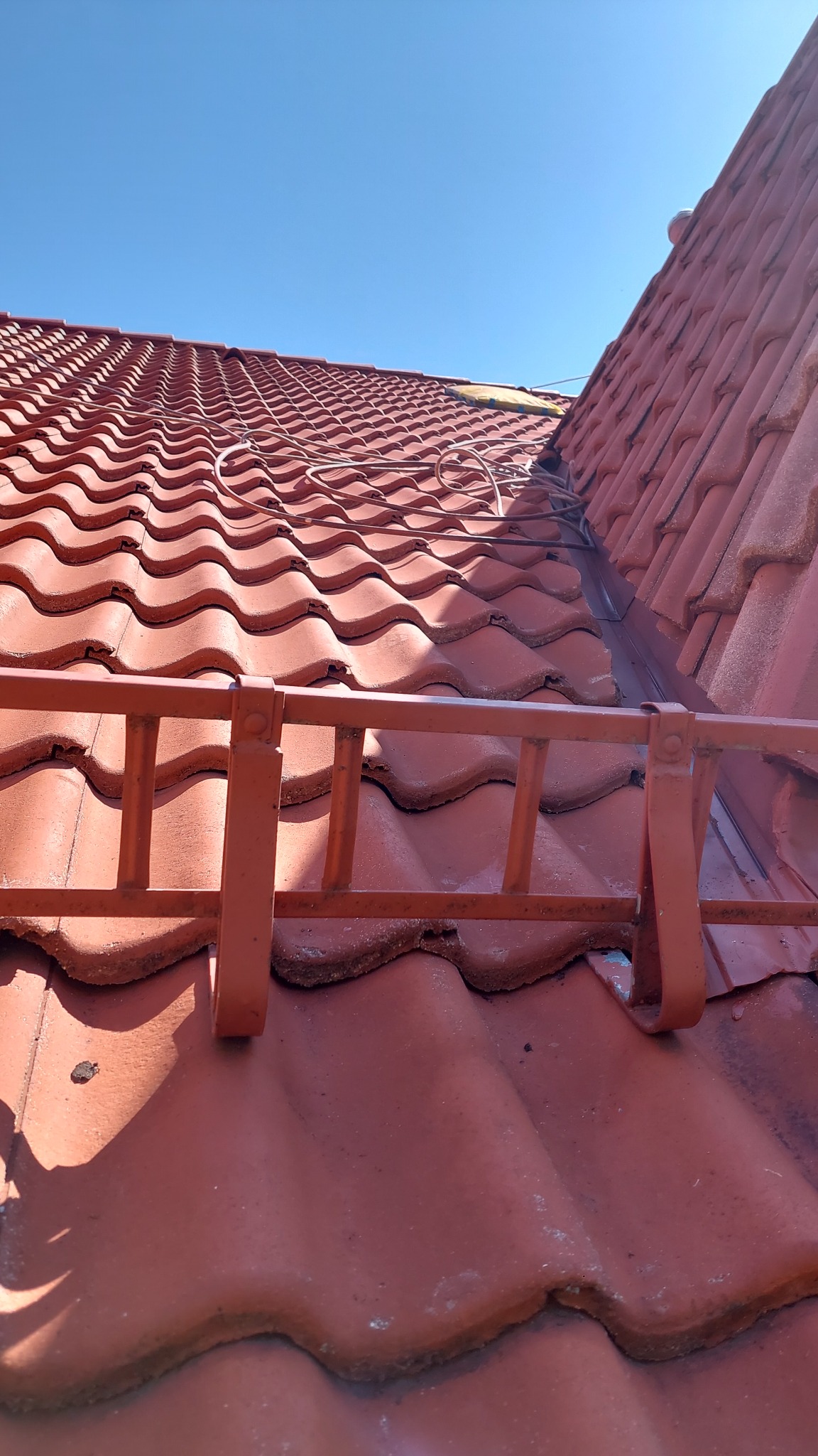 Czerwona dachówka ceramiczna, zabezpieczona metalową barierką przeciwśniegową, widziana pod kątem w słoneczny dzień. Na dachu leżą przewody.
