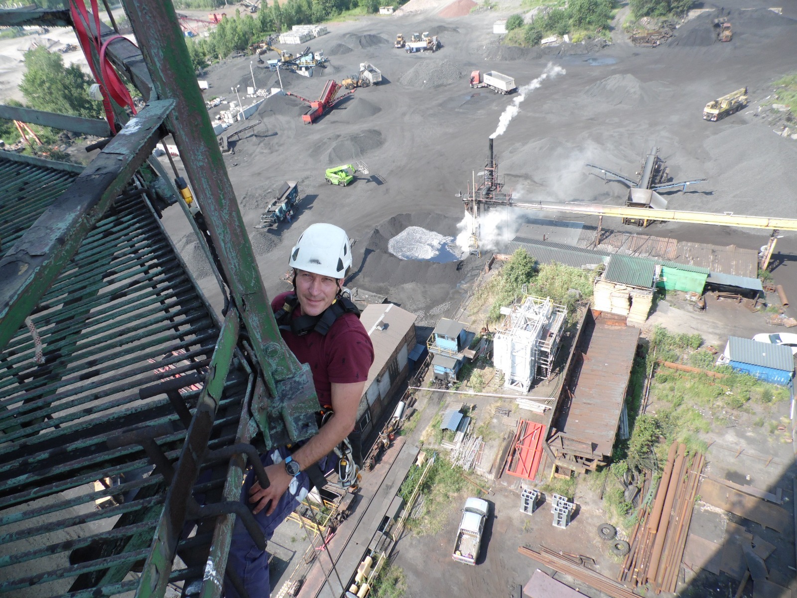 Pracownik w kasku, na drabinie konstrukcji przemysłowej, z widokiem na teren zakładu z hałdami materiałów i kominem. Ujęcie z góry, oświetlenie dzienne.
