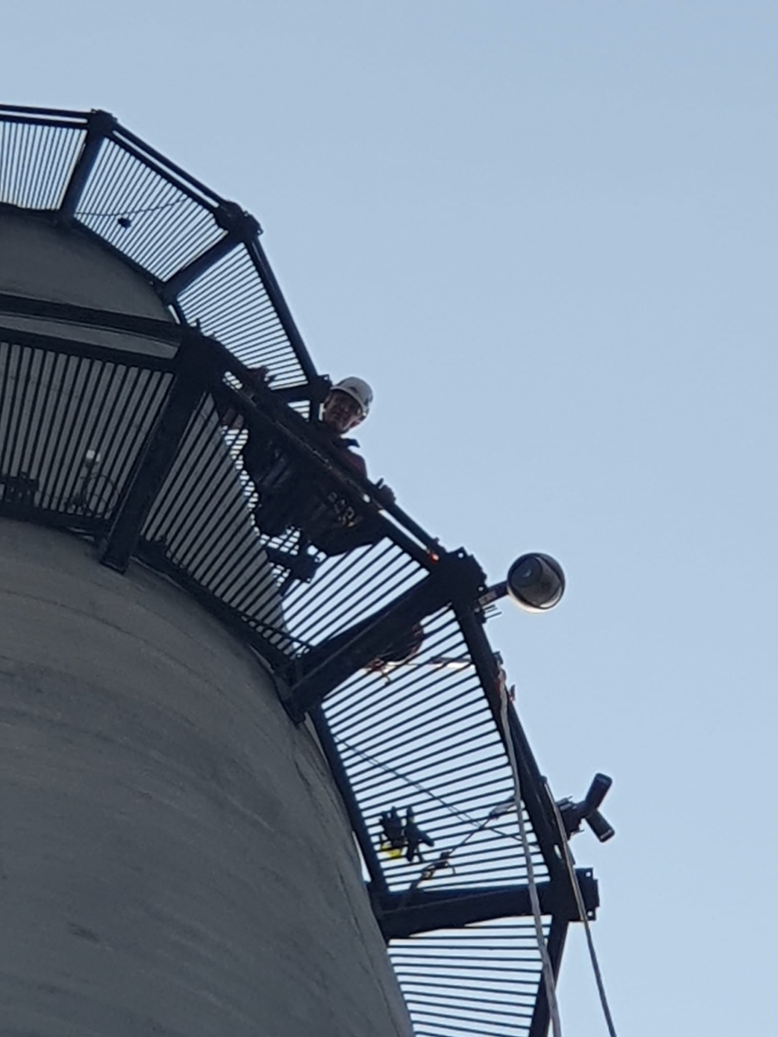 Pracownik w kasku na szczycie betonowej konstrukcji z metalową klatką, zabezpieczony linami, wykonuje prace na wysokości, widok z dołu.