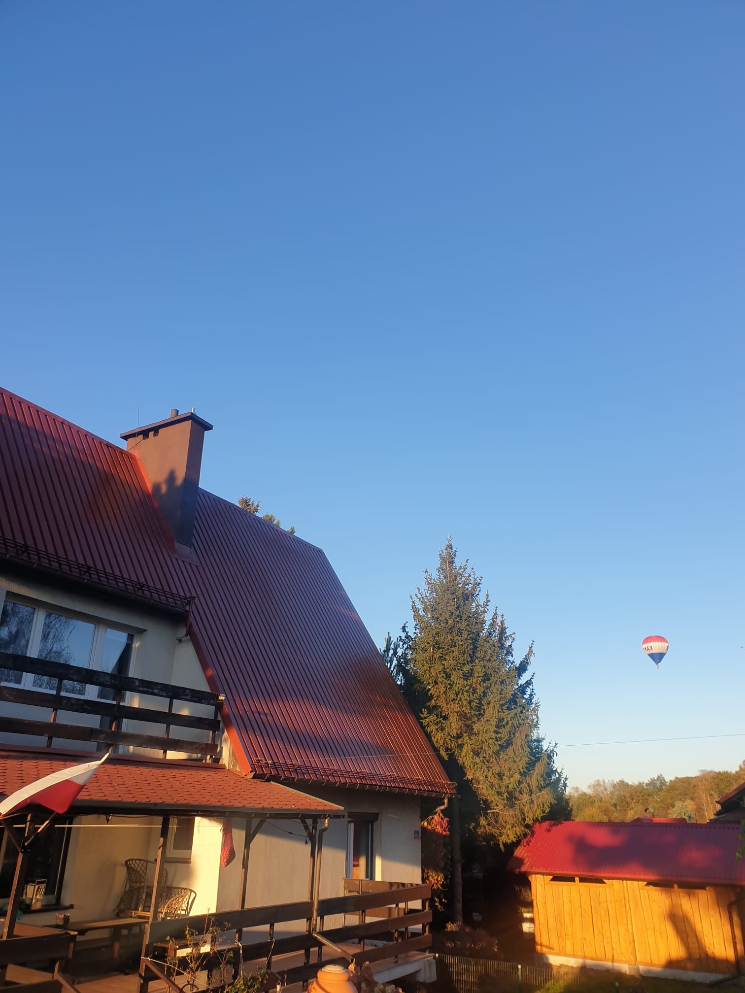 Dom z czerwonym dachem w promieniach słońca, wysoka choinka obok, na błękitnym niebie balon. Widok z balkonu.
