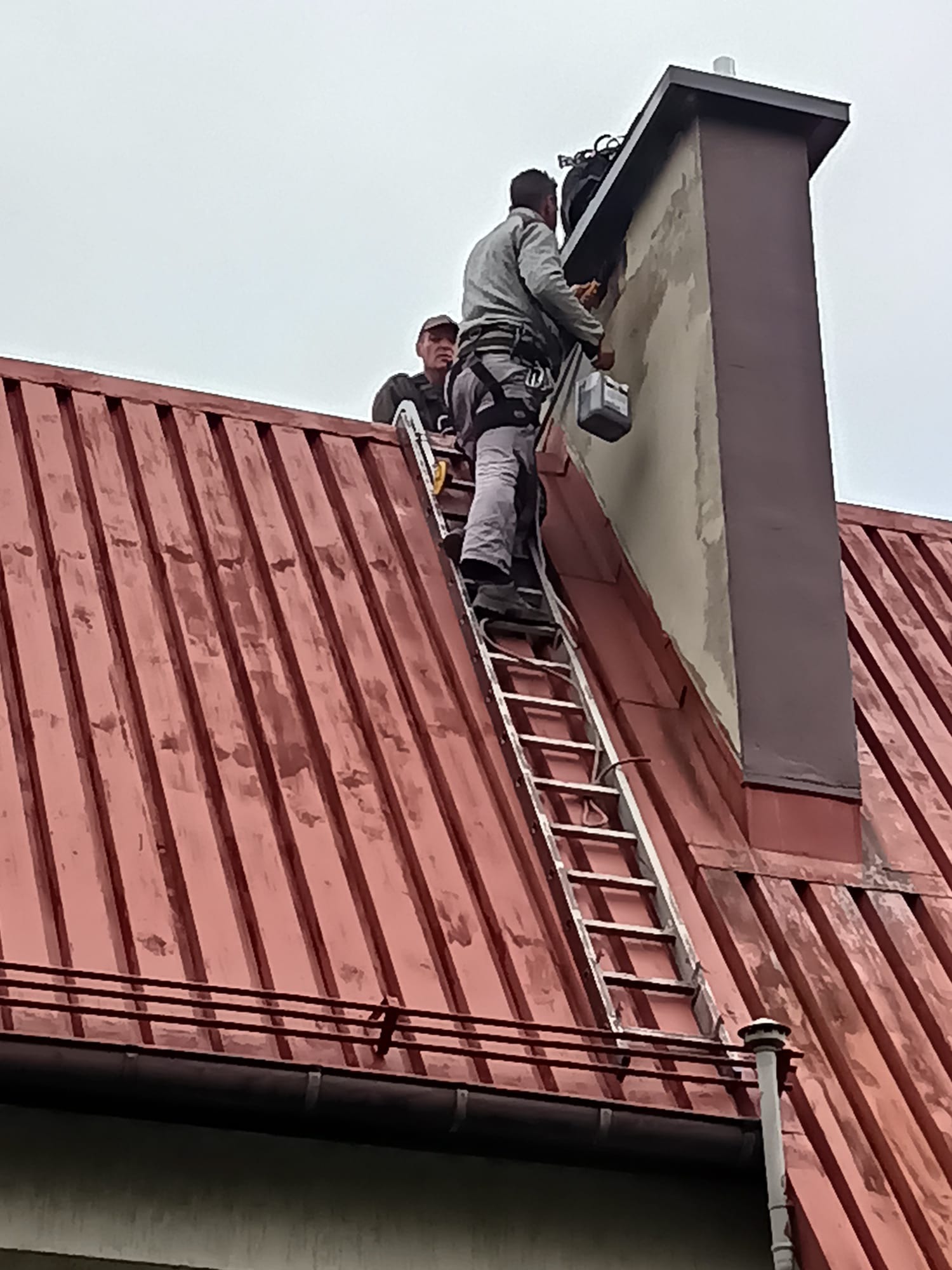 Pracownik na drabinie, w uprzęży, maluje komin na czerwonym dachu. Drugi pracownik asekuruje. Widok z dołu, pochmurne niebo.
